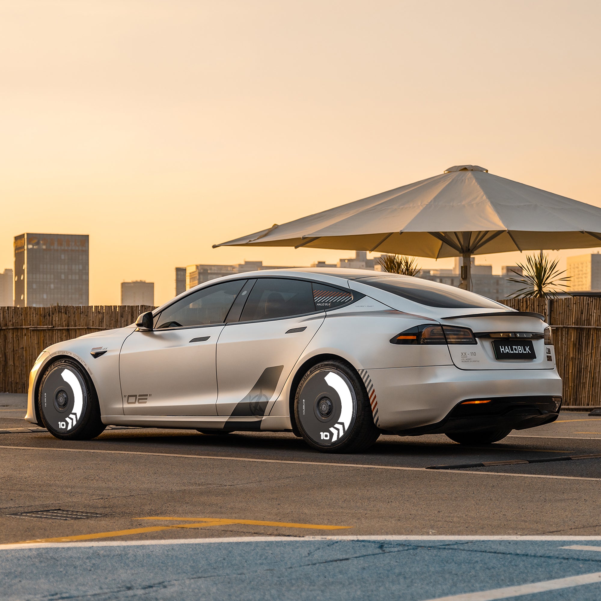 A Tesla Model S featuring HALODISC 2 Arrow Wheel Covers from HALOBLK Store is parked in a modern city at sunset, with bold wheels, buildings, and a large sun umbrella in the background.