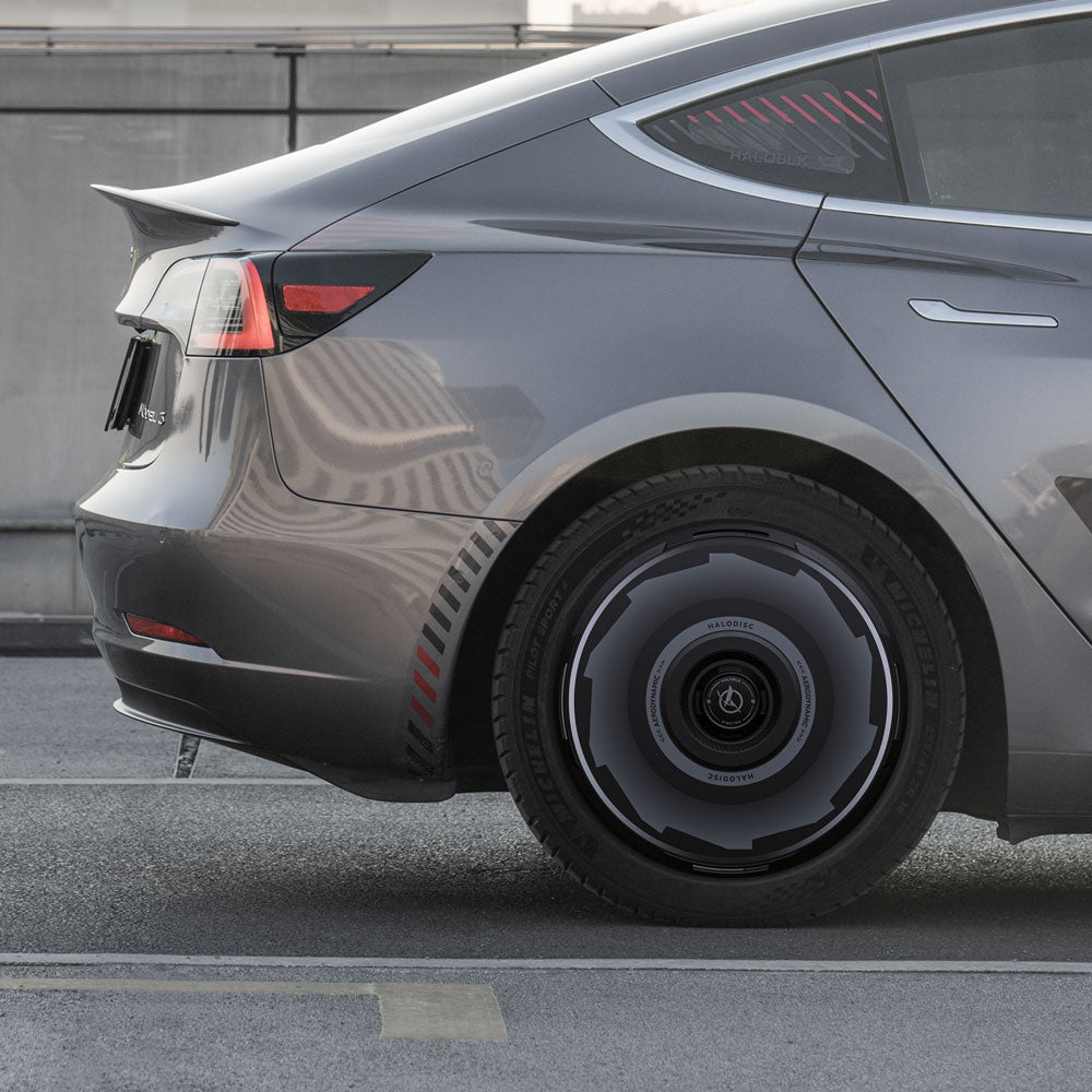 A close-up side view of a Tesla Model 3 (2019-2023) with HALODISC 2 Wheel Covers by HALOBLK Store on its 19" Zero-G wheels, featuring a futuristic wheel design and red accents, parked on an urban street.