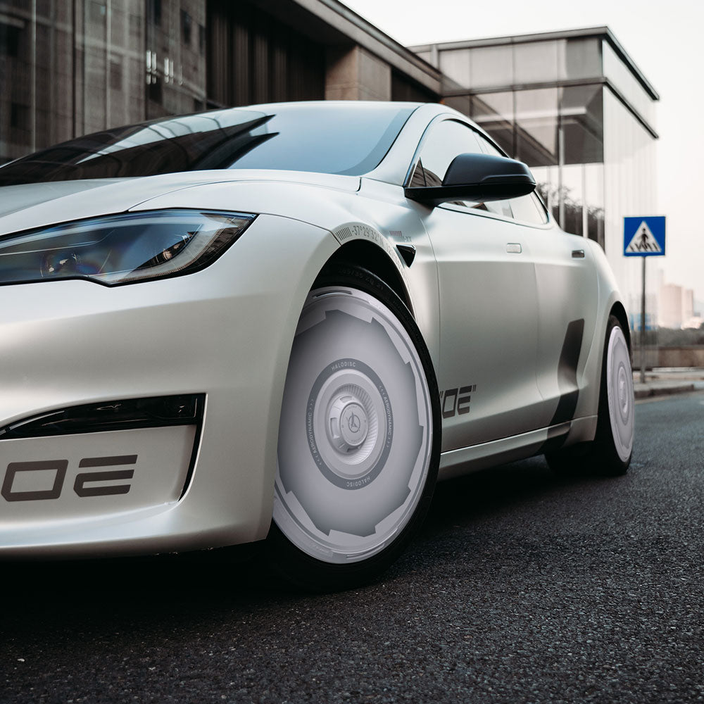 A Tesla Model S with HALODISC 2 Wheel Covers by HALOBLK Store is parked on a city street near a glass building, featuring stylish 21" Arachnid Vision-GT rims. A pedestrian crossing sign appears in the background.