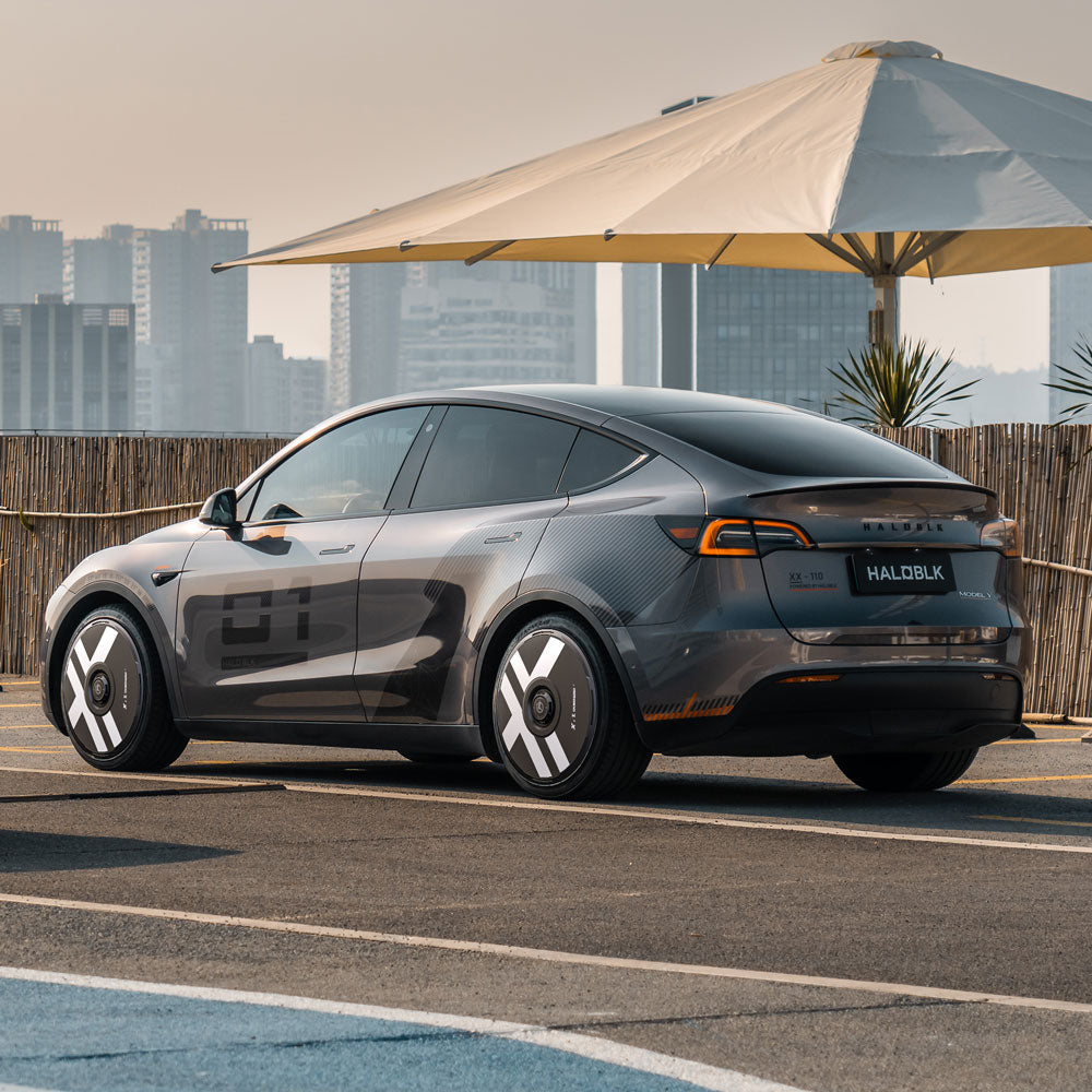 A Tesla Model Y with HALODISC 2 Wheel Covers by HALOBLK Store is parked on a rooftop lot near a wooden fence, city buildings in the background, and a large white sun umbrella nearby.
