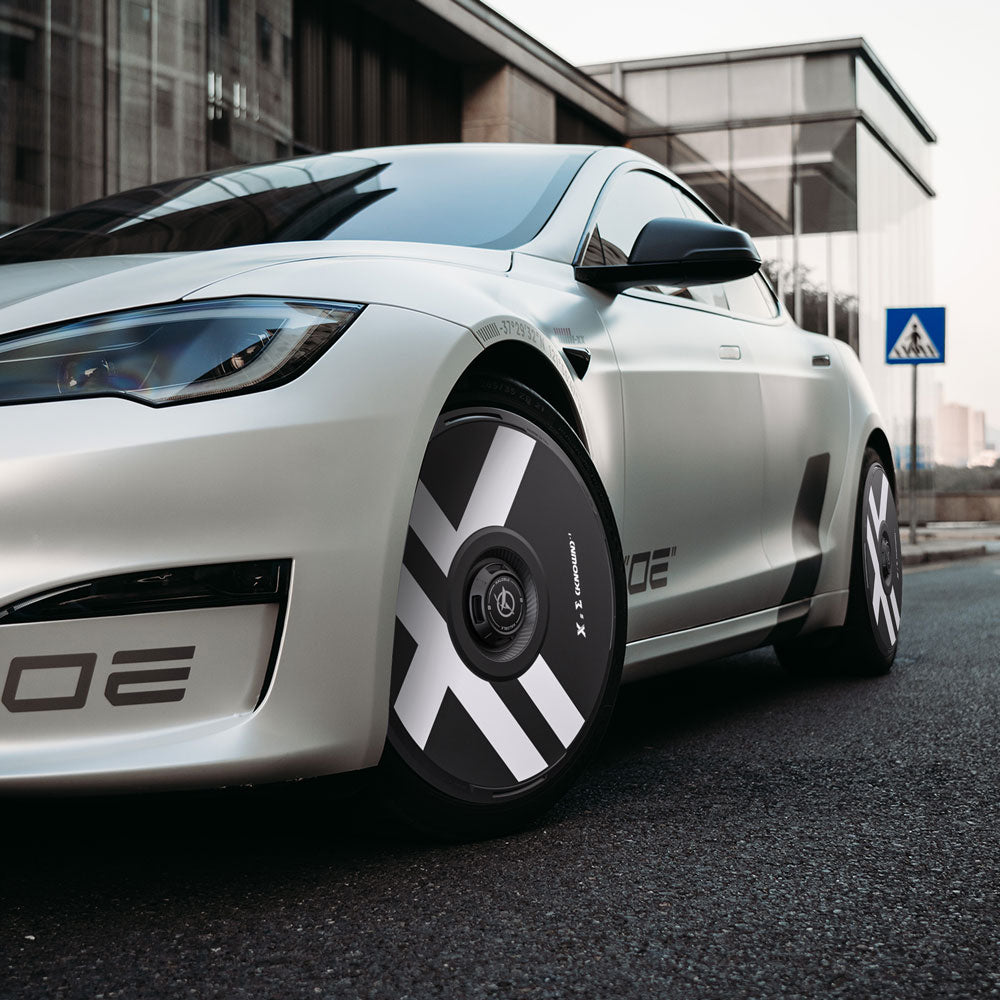 A sleek white Tesla Model S with HALODISC 2 Wheel Covers for 21" Arachnid wheels by HALOBLK Store, sporting bold black and white geometric patterns, parked on a street near a glass building and a crosswalk sign.