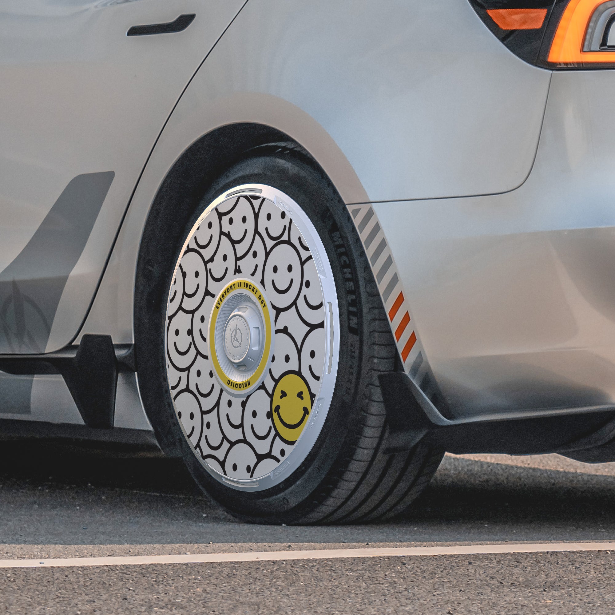 A close-up of a Tesla Model 3 Highland 19" Nova Wheel fitted with HALODISC 2 Wheel Covers from HALOBLK Store, featuring black and white smiley faces and one yellow smiley. The car is parked on asphalt.