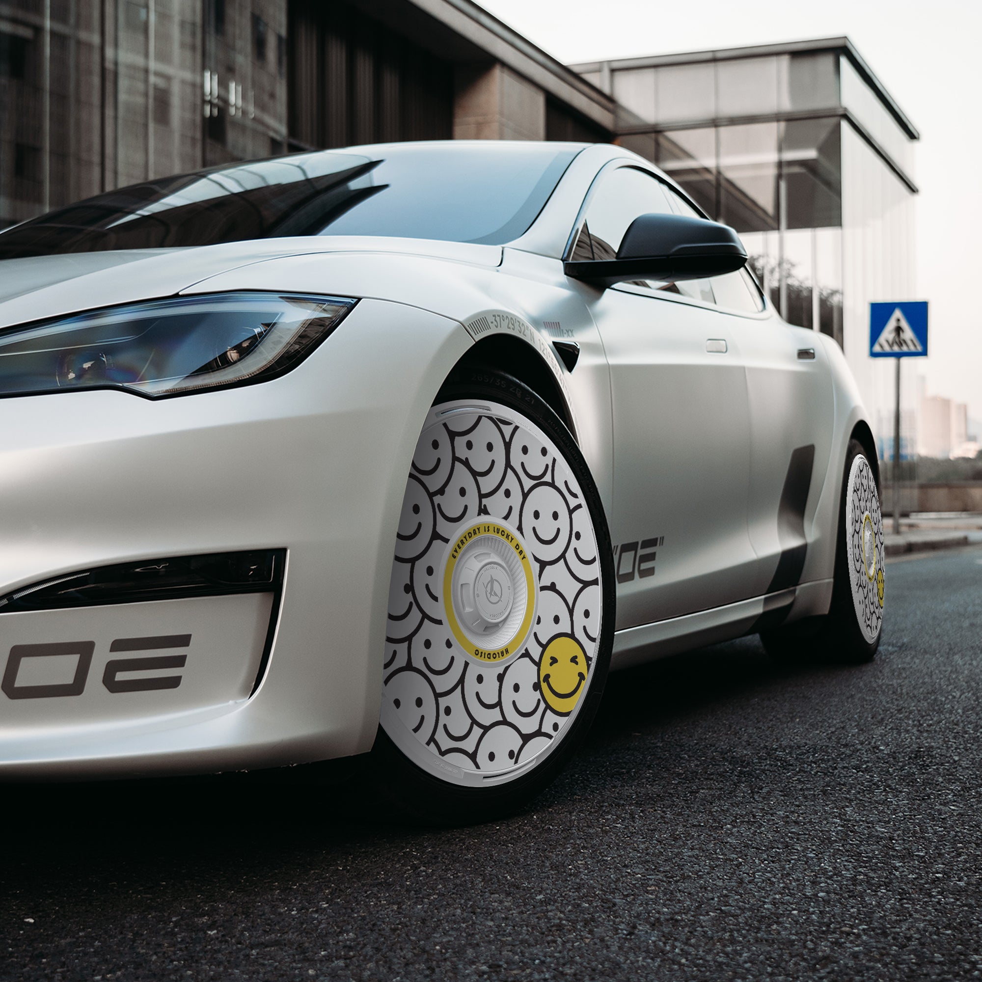 A white Tesla Model S with HALODISC 2 Wheel Covers (“Lucky Day” by HALOBLK Store) featuring black-outlined and one yellow smiley face is parked on the street, with a modern building and crosswalk sign in the background.