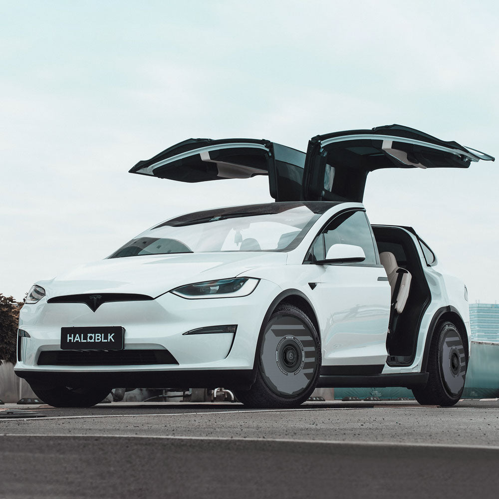 A white Tesla Model X (2021-2025) with HALODISC 2 Wheel Covers by HALOBLK Store is parked outdoors under a cloudy sky, its falcon-wing doors open and a front license plate reading HALOBLK.