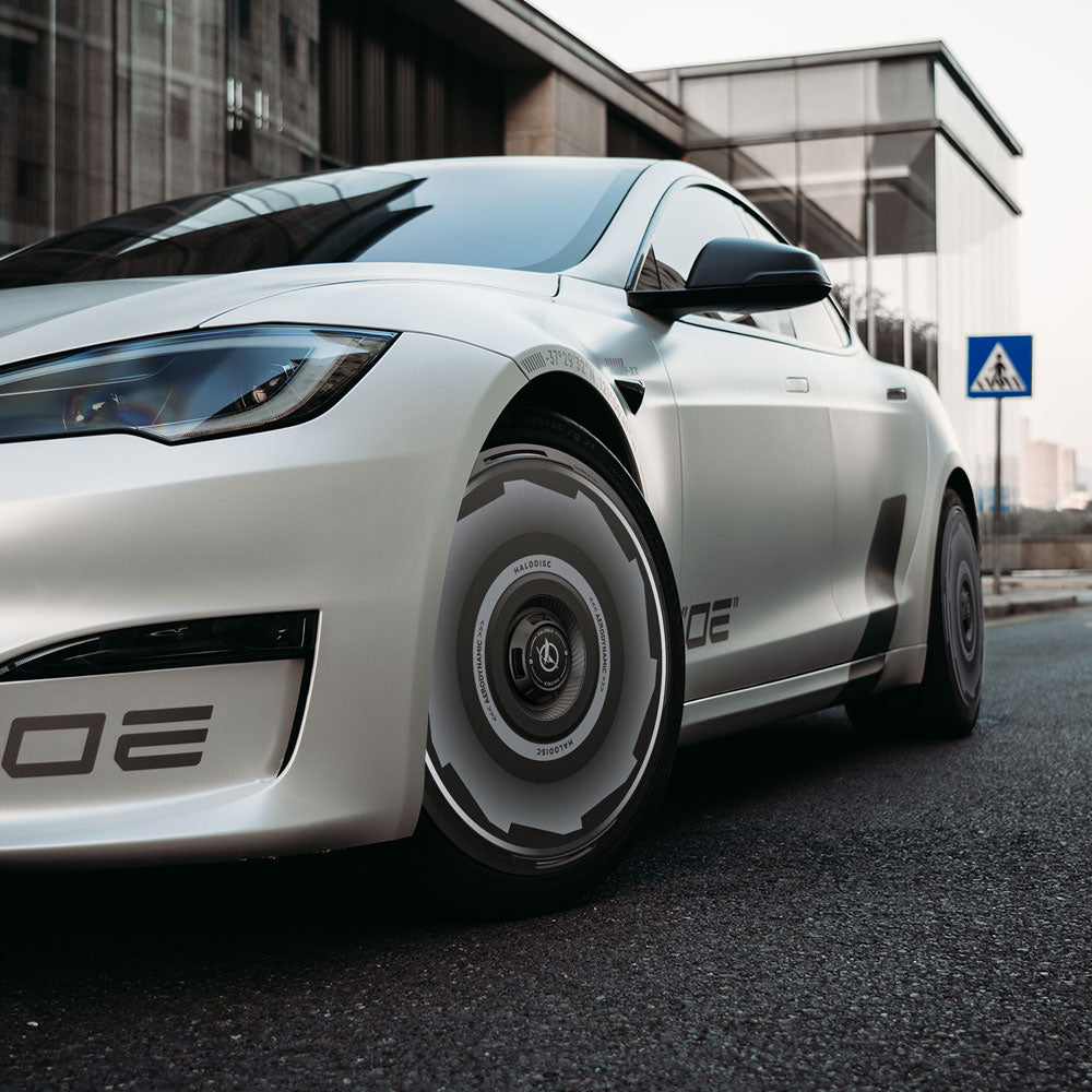 A white Tesla Model S with HALODISC 2 Vision-GT wheel covers (19" Magnetite, HALOBLK Store) is parked on a street by a glass building and a pedestrian crossing sign.