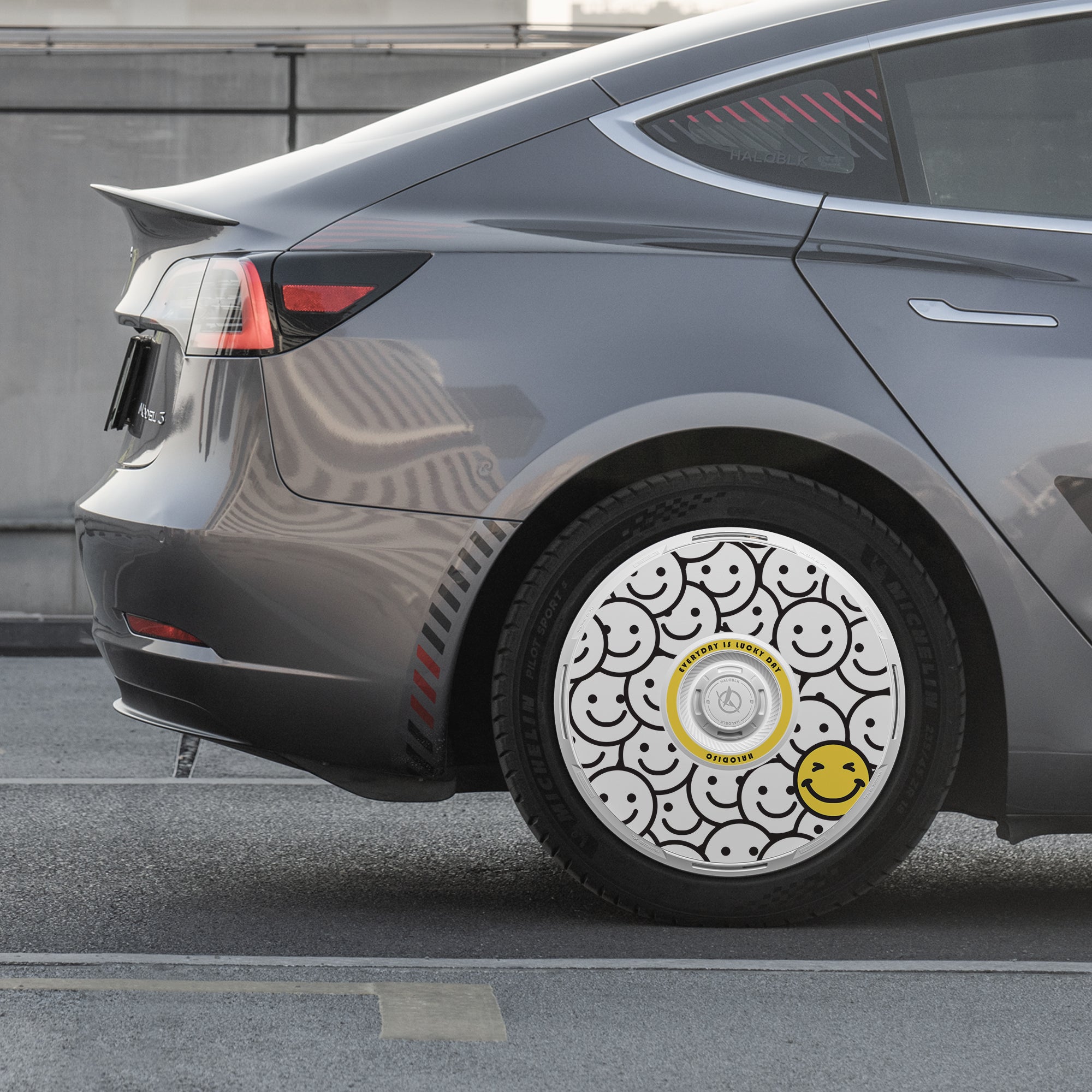A gray car parked on the street has HALODISC 2 "Lucky Day" wheel covers by HALOBLK Store for Tesla Model 3 (2017-2018), featuring a pattern of black outlined smiley faces and one yellow smiley face on the rear wheel.