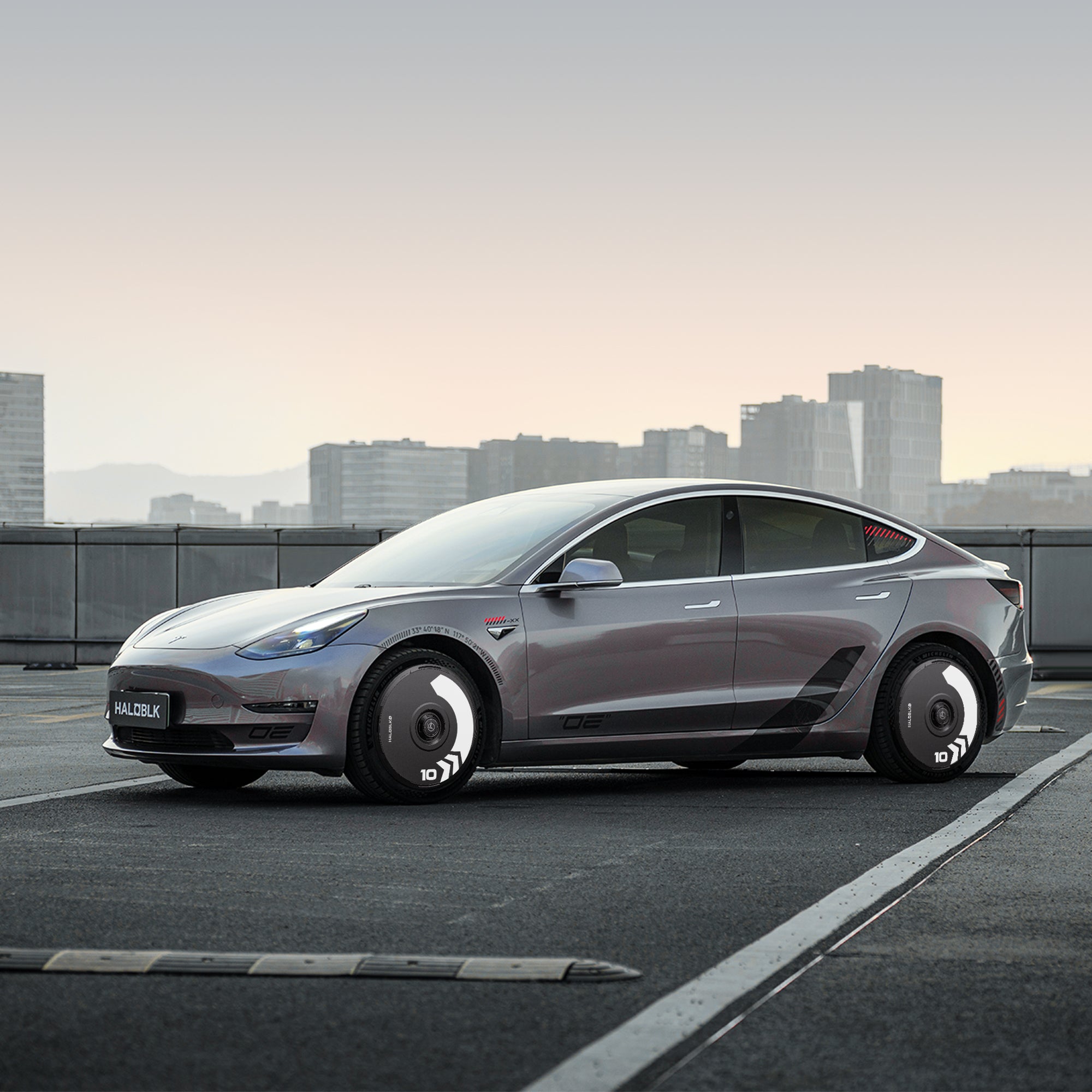 A silver Tesla Model 3 (2018-2023) with HALODISC 2 Wheel Covers by HALOBLK Store on 19" Stiletto Wheels is parked on a rooftop against a city skyline at dusk, featuring an advanced, sleek look and futuristic wheel design.