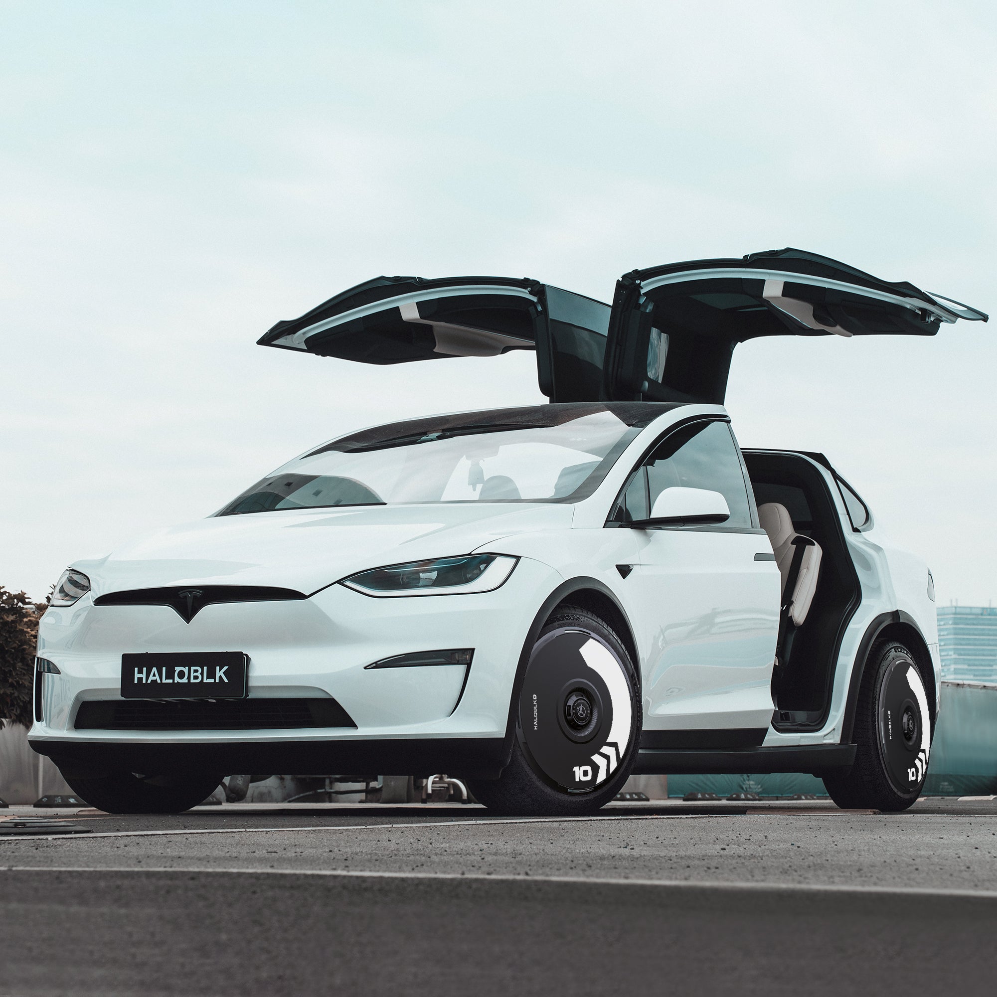 A white Tesla Model X (2016-2020) with gull-wing doors open is parked outdoors under an overcast sky, featuring HALODISC 2 Arrow wheel covers by HALOBLK Store on its 20" Slipstream wheels. The front plate reads 'HALOBLK.'.