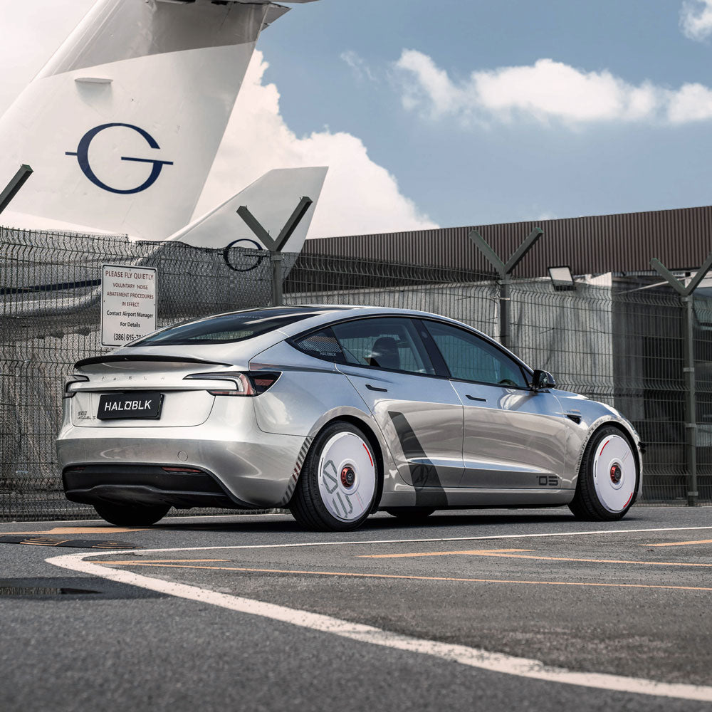 A silver Tesla sedan with HALODISC 2 Wheel Covers (custom numbers, white) from HALOBLK Store is parked near an airport runway; a large airplane and fence are visible under partly cloudy skies. The license plate reads “HALD8L8K.”.