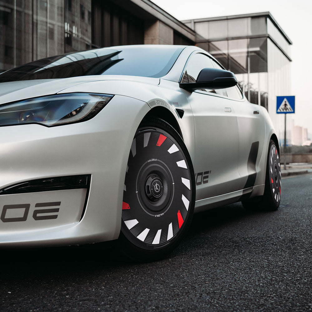 A sleek silver Tesla Model S with HALODISC 2 Wheel Covers by HALOBLK Store—featuring black, red, and white patterns—sits parked near a modern glass building by a pedestrian crossing sign.