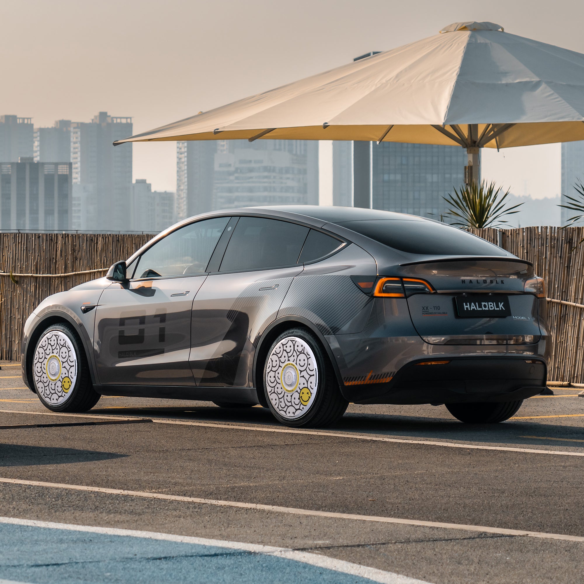 A Tesla Model Y with HALODISC 2 "Lucky Day" wheel covers from HALOBLK Store is parked outdoors on a sunny day near a large umbrella and bamboo fencing, with a city skyline in the background.
