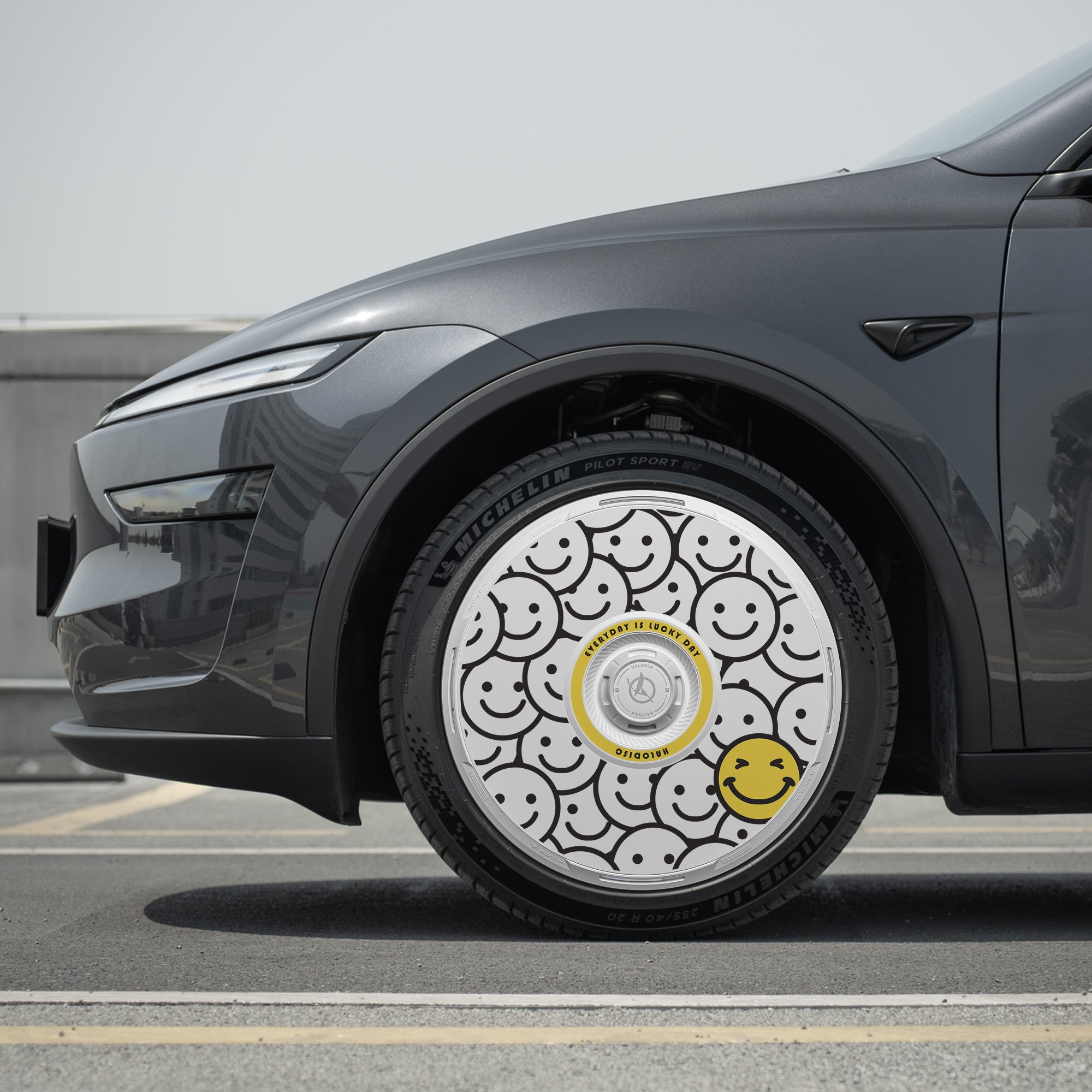 A close-up of a Tesla Model Y’s Juniper 19" Crossflow wheel with a HALODISC 2 Wheel Cover “Lucky Day” from HALOBLK Store, featuring black-outlined smiley faces on white and a large yellow smiley near the bottom right.