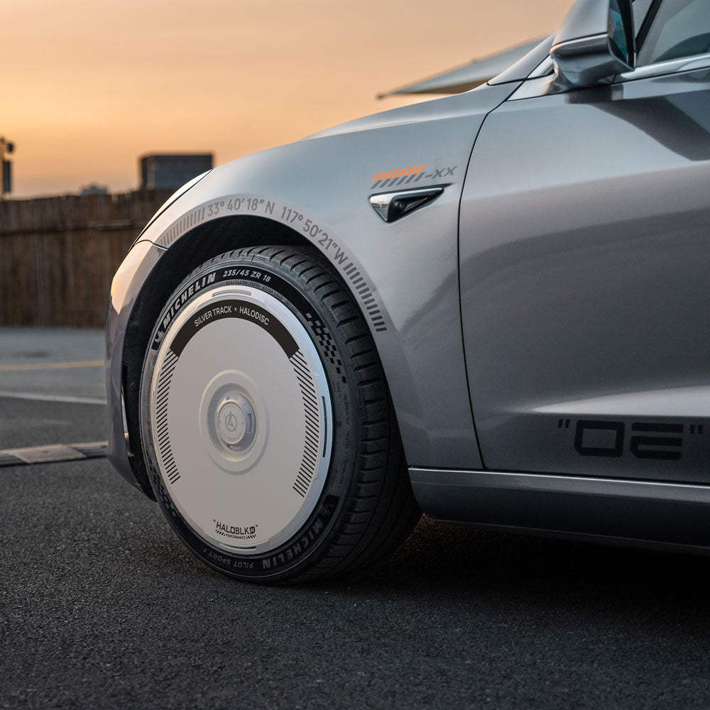 Close-up of a Tesla Model 3 (2017-2018) with HALODISC 2 Silver Track 19" wheel covers by HALOBLK Store, showing detailed futuristic design, parked on pavement at sunset with a wooden fence in the background.