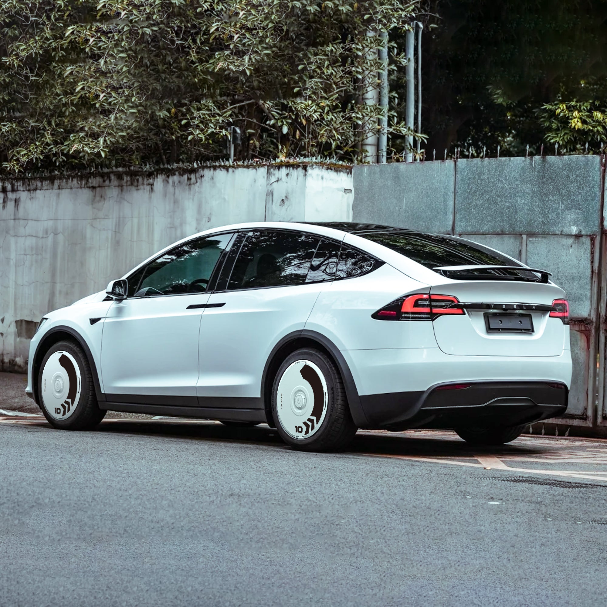 A white Tesla Model X with black trim featuring HALODISC 2 Wheel Covers for 2016-2020 20" Slipstream Wheels from HALOBLK Store is parked beside a tall concrete wall and green foliage.