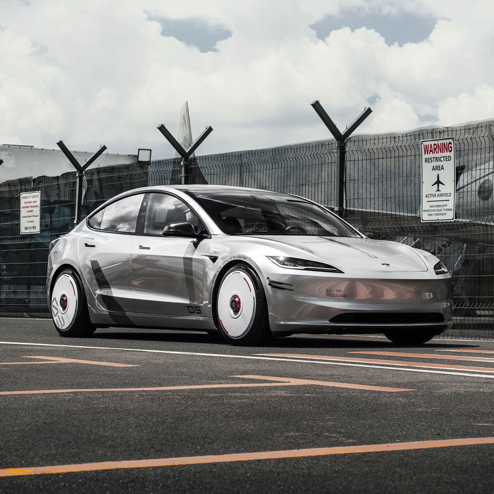 A shiny silver Tesla with HALODISC 2 Wheel Covers for Tesla (Custom Numbers - White) from HALOBLK Store is parked in an industrial area beside a chain-link fence with barbed wire and warning signs under a partly cloudy sky.