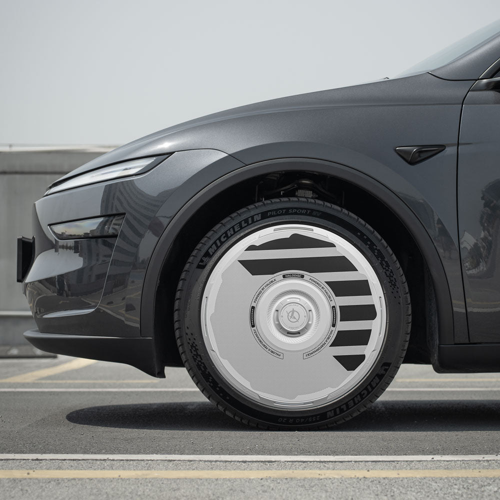 Close-up of the front left wheel of a dark gray car fitted with HALODISC 2 Wheel Covers for Tesla Model Y Juniper 19" Crossflow Wheels by HALOBLK Store, showing unique silver and black accents on a city rooftop under a clear sky.