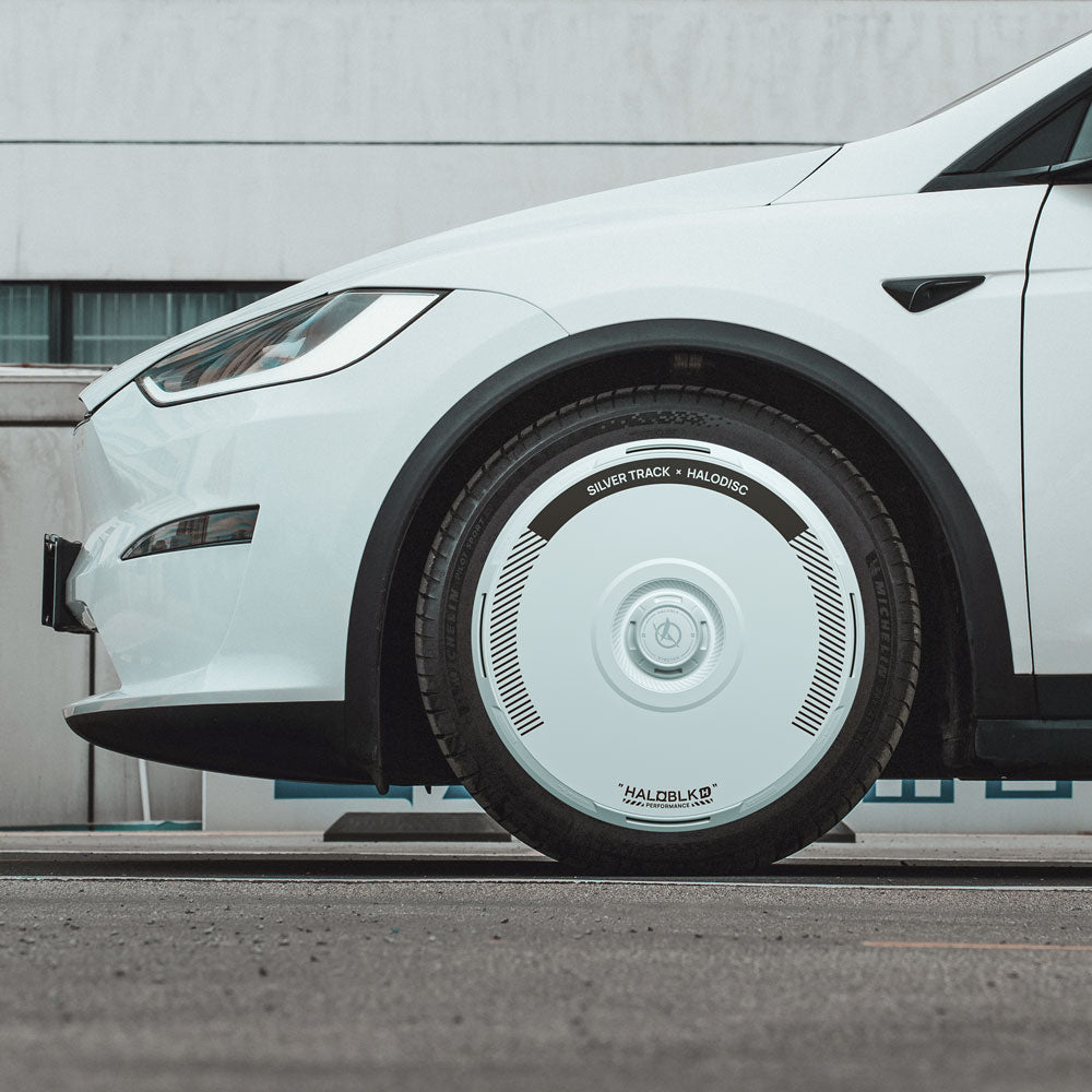 A close-up side view of a white Tesla Model X (2021–2025) shows a shiny, circular HALODISC 2 Silver Track wheel cover (HALOBLK Store) on a 20" Cyberstream wheel, parked on asphalt with a modern building in the background.