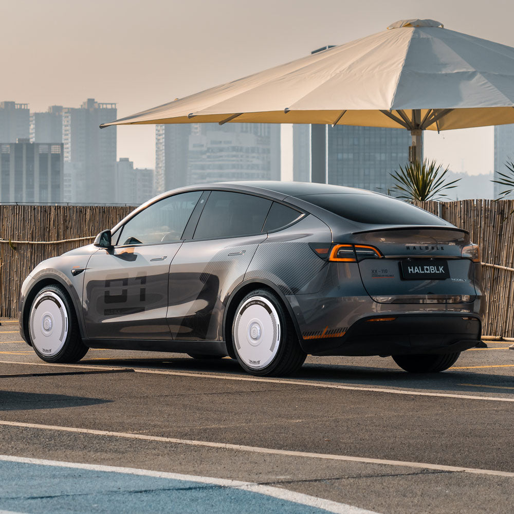 A gray Tesla Model Y with HALODISC 2 Silver Track wheel covers from HALOBLK Store is parked outdoors, set against a city skyline, bamboo fence, and a large patio umbrella.