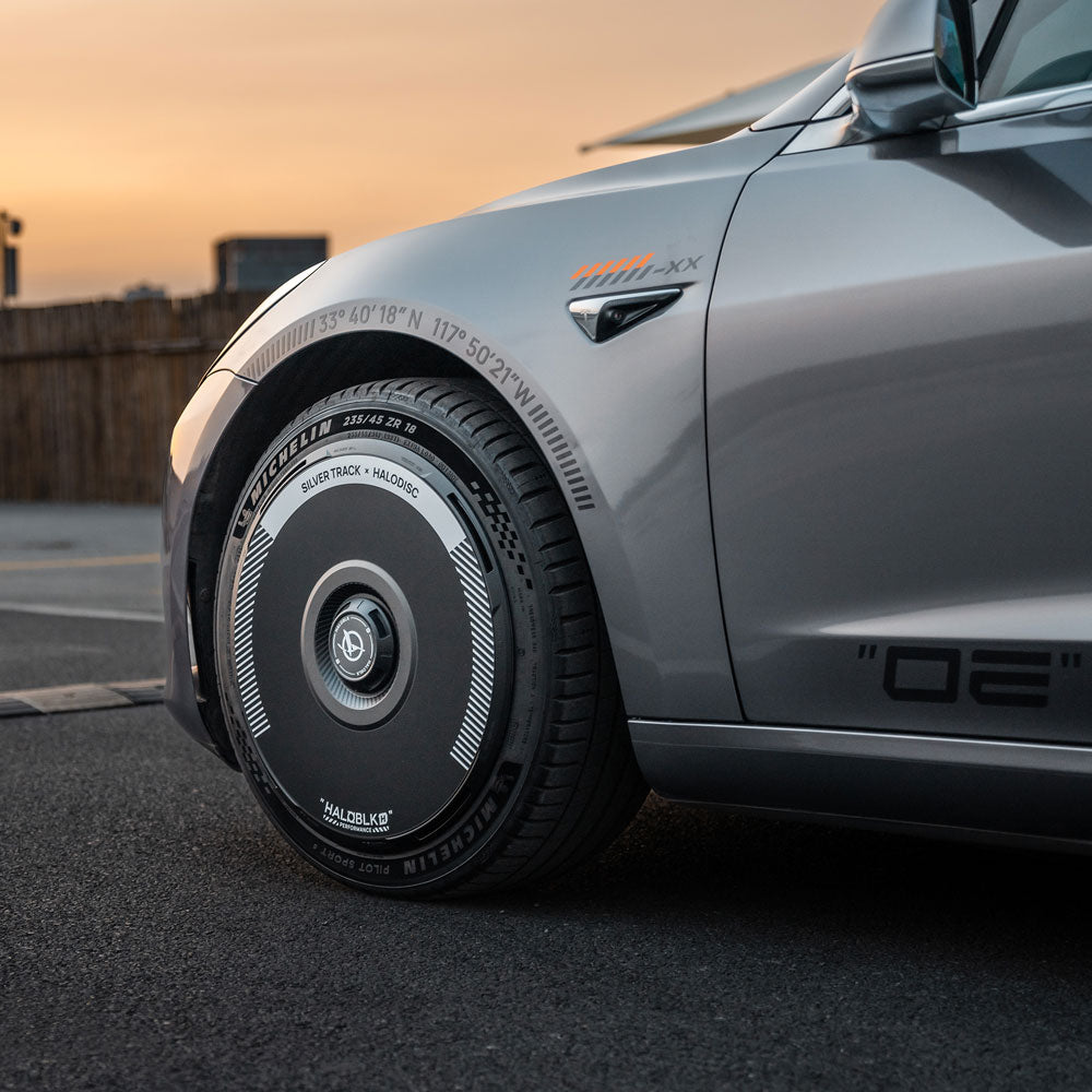 Close-up of HALODISC 2 Silver Track 19" Wheel Cover by HALOBLK Store on a Tesla Model 3 (2017-2018), showing its futuristic design and tire tread, parked on asphalt with a sunset sky in the background.