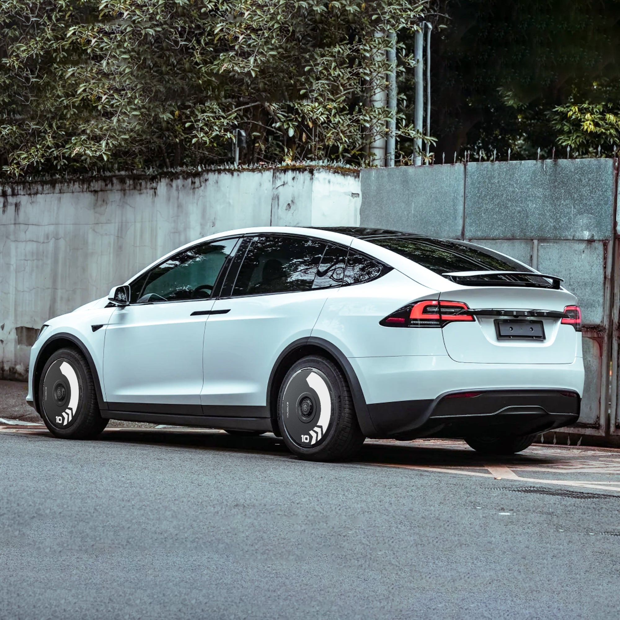 A white Tesla Model X parked by a concrete wall features HALODISC 2 Wheel Covers for 2021-2025 20" Perihelix Wheels (Arrow) by HALOBLK Store, showcasing black trim and eye-catching spiral-patterned covers.