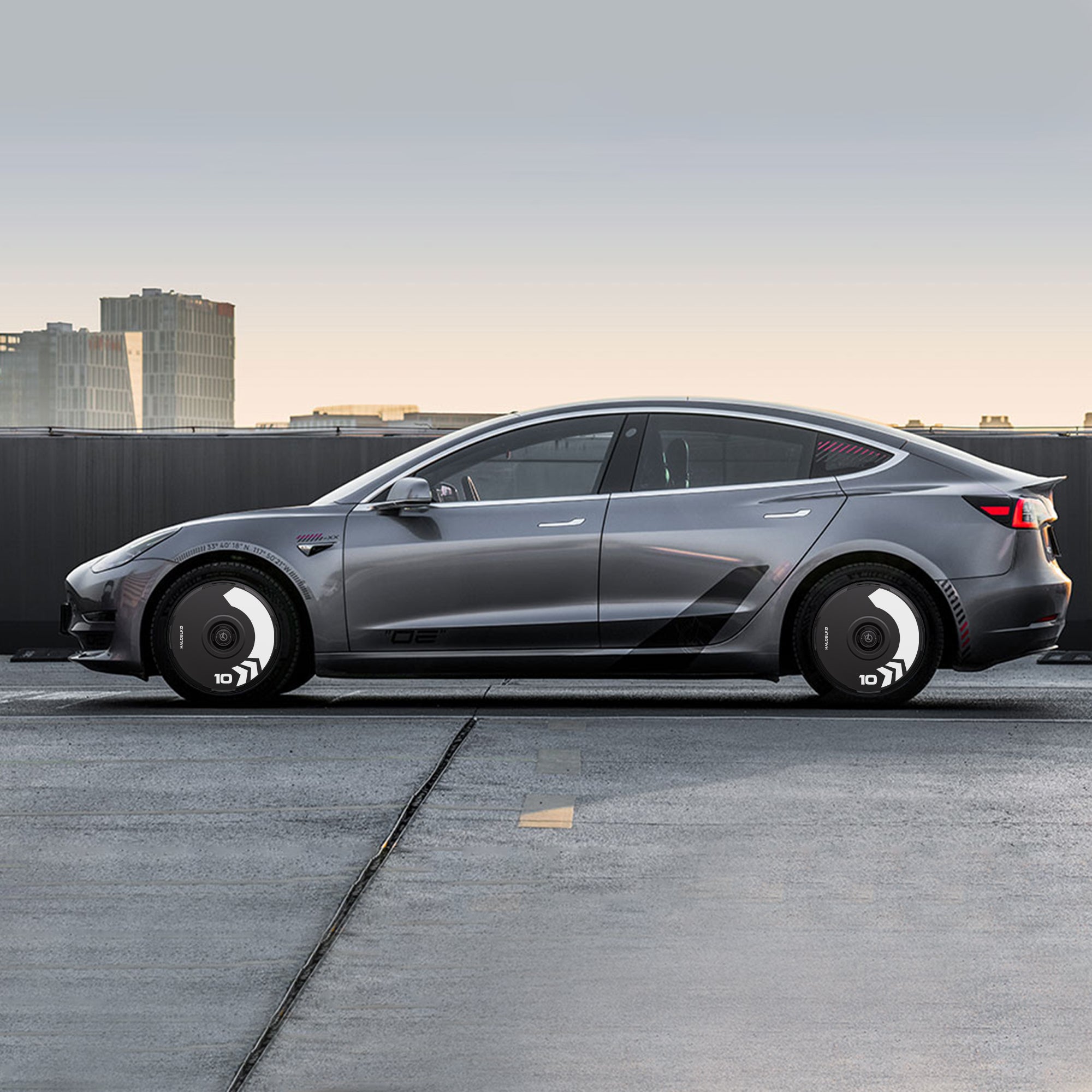 A sleek gray Tesla Model 3 with HALODISC 2 Wheel Covers by HALOBLK Store is parked on a rooftop, its 19" Stiletto Wheels featuring bold, arrow-patterned black and white hubcaps against a modern city backdrop.