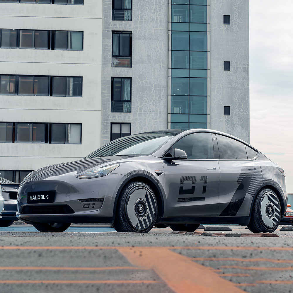A silver Tesla Model Y with HALODISC 2 Wheel Covers from HALOBLK Store, featuring custom graphics, black accents, and the number 01 on the sides, is parked in front of a modern multi-story building.