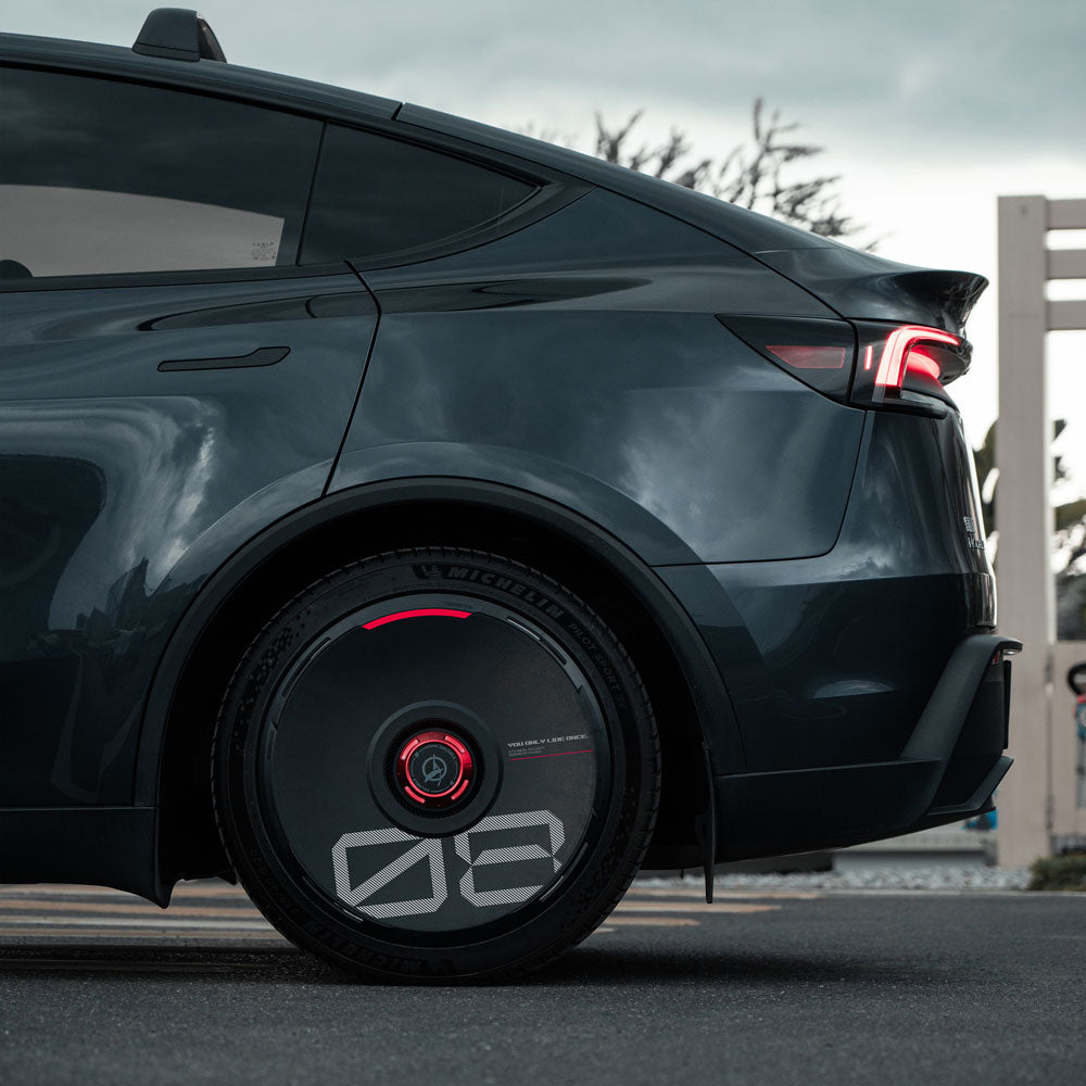 A close-up side view of a dark gray Tesla showcases a custom HALODISC 2 Wheel Cover for Tesla by HALOBLK Store, featuring black and red accents with bold white numbers. The car is parked outdoors on a cloudy day.