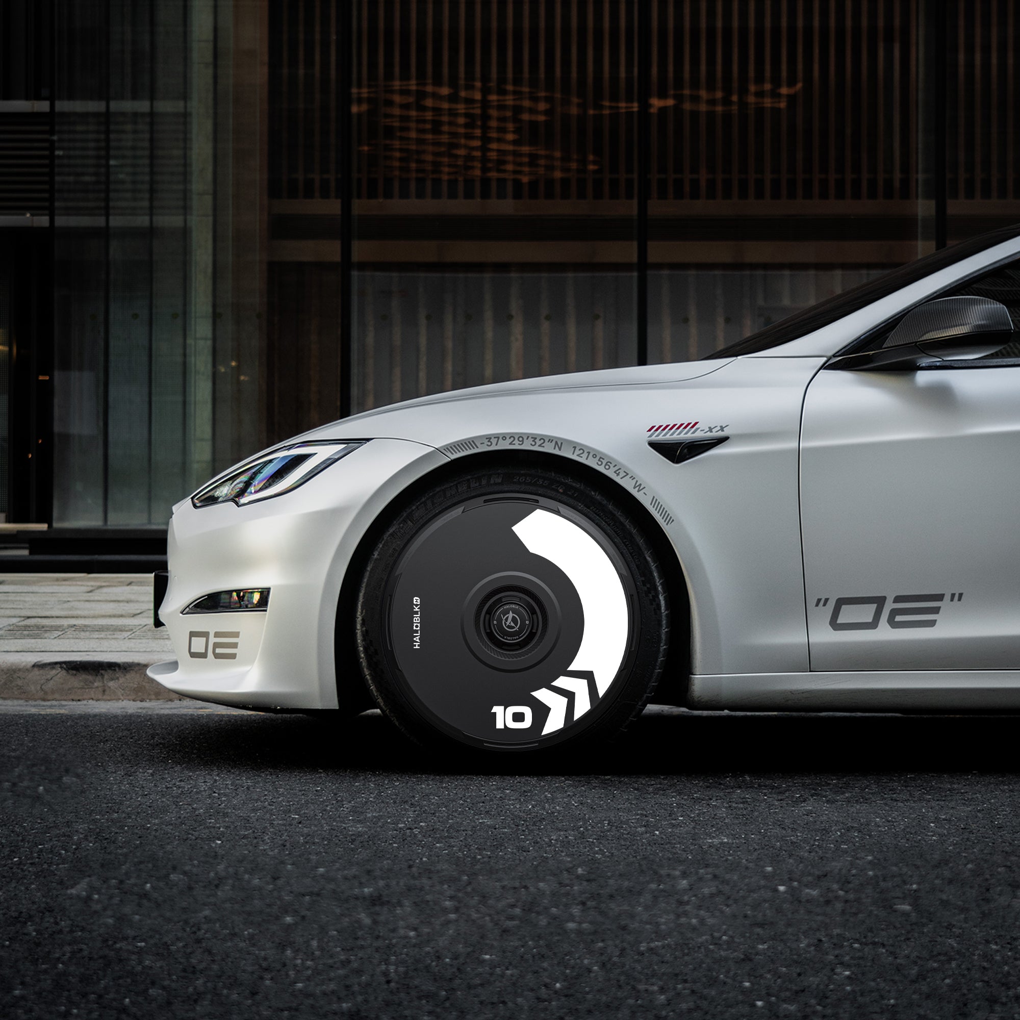 A white sports car parked on a city street features HALODISC 2 Arrow Wheel Covers from HALOBLK Store for Tesla Model S 19" Slipstream wheels, with bold OE decals and black wheels accented by white geometric patterns and the number 10.