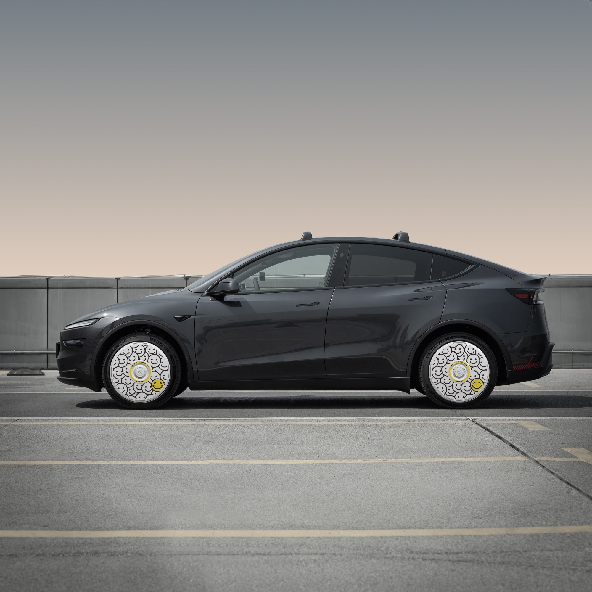 A black car with HALODISC 2 Wheel Covers for Tesla Model Y Juniper 19" Crossflow Wheels (Lucky Day) by HALOBLK Store sits on a rooftop, showcasing artistic white, gray, and yellow designs against a concrete barrier and gradient sky.
