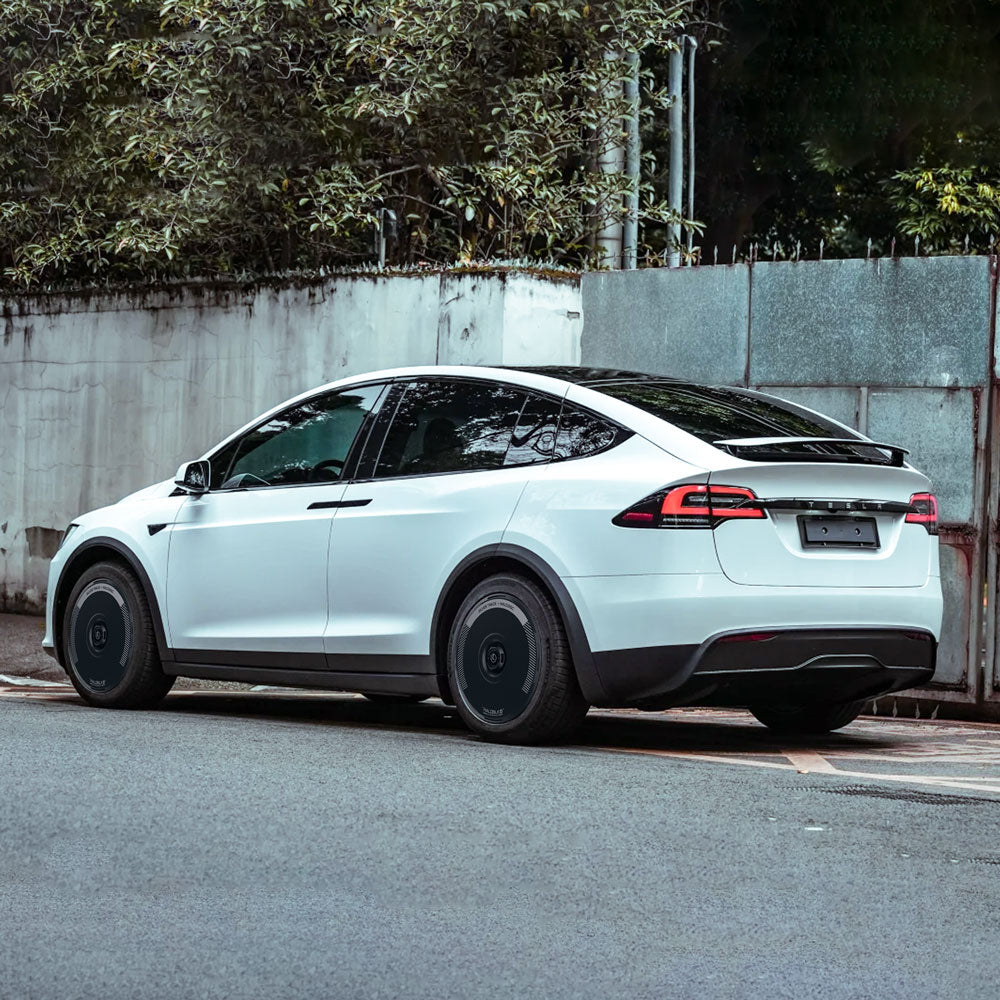 A white Tesla Model X (2021-2025) with HALODISC 2 Silver Track 20" wheel covers from HALOBLK Store is parked on a street by a sidewalk, with trees and a tall light wall in the background, viewed from the rear side.