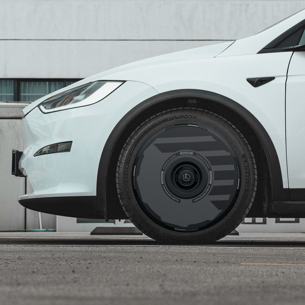 Side view of a white Tesla Model X (2016-2020) parked on a street, highlighting the front wheel with HALODISC 2 Wheel Covers from HALOBLK Store, featuring a dark, aerodynamic rim design; part of a white building is visible in the background.