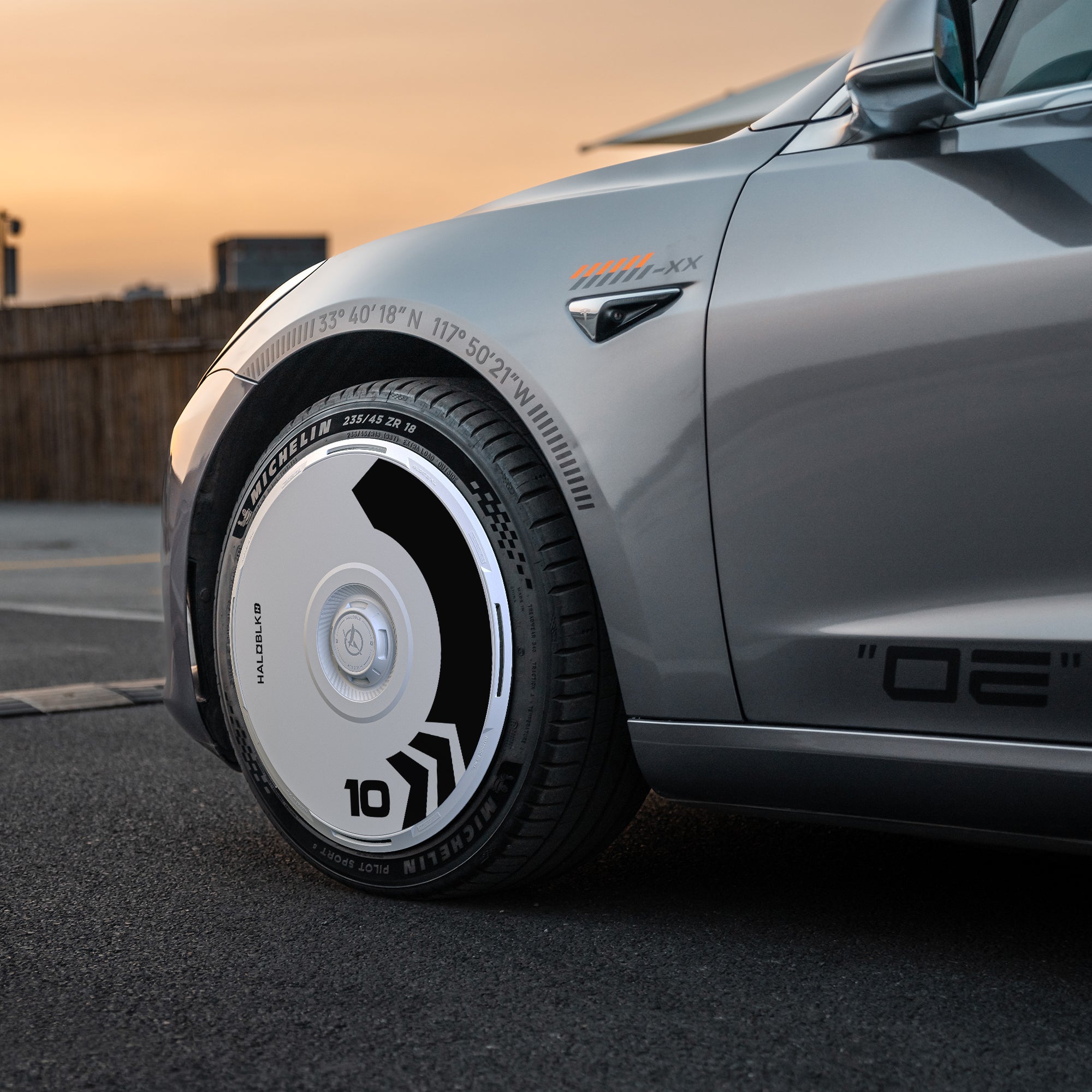 Close-up of a Tesla Model 3 (2019-2023) front wheel with HALOBLK Store’s HALODISC 2 Arrow wheel cover, featuring black and white geometric designs and "10," fitted on a Michelin tire, parked on a street at sunset.
