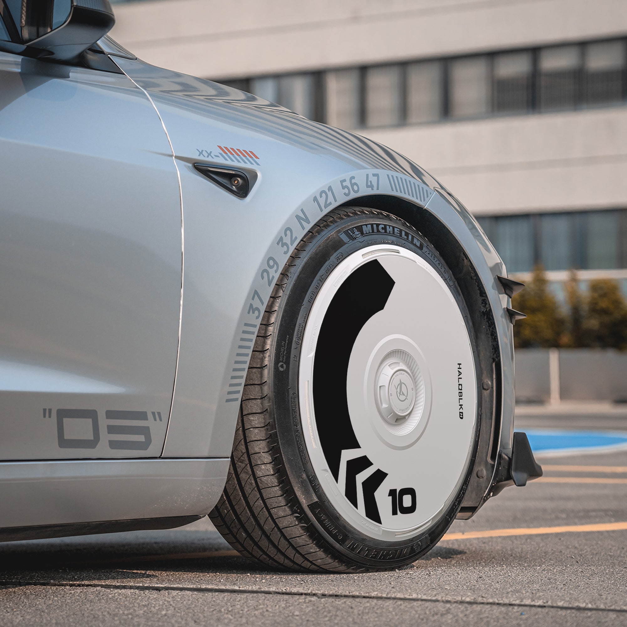 Close-up of HALODISC 2 Wheel Covers for Tesla Model 3 Highland 20" Warp Wheels by HALOBLK Store—futuristic silver car with unique white disk-like hubcap, “10” mark, black geometric patterns, and a modern building in the background.