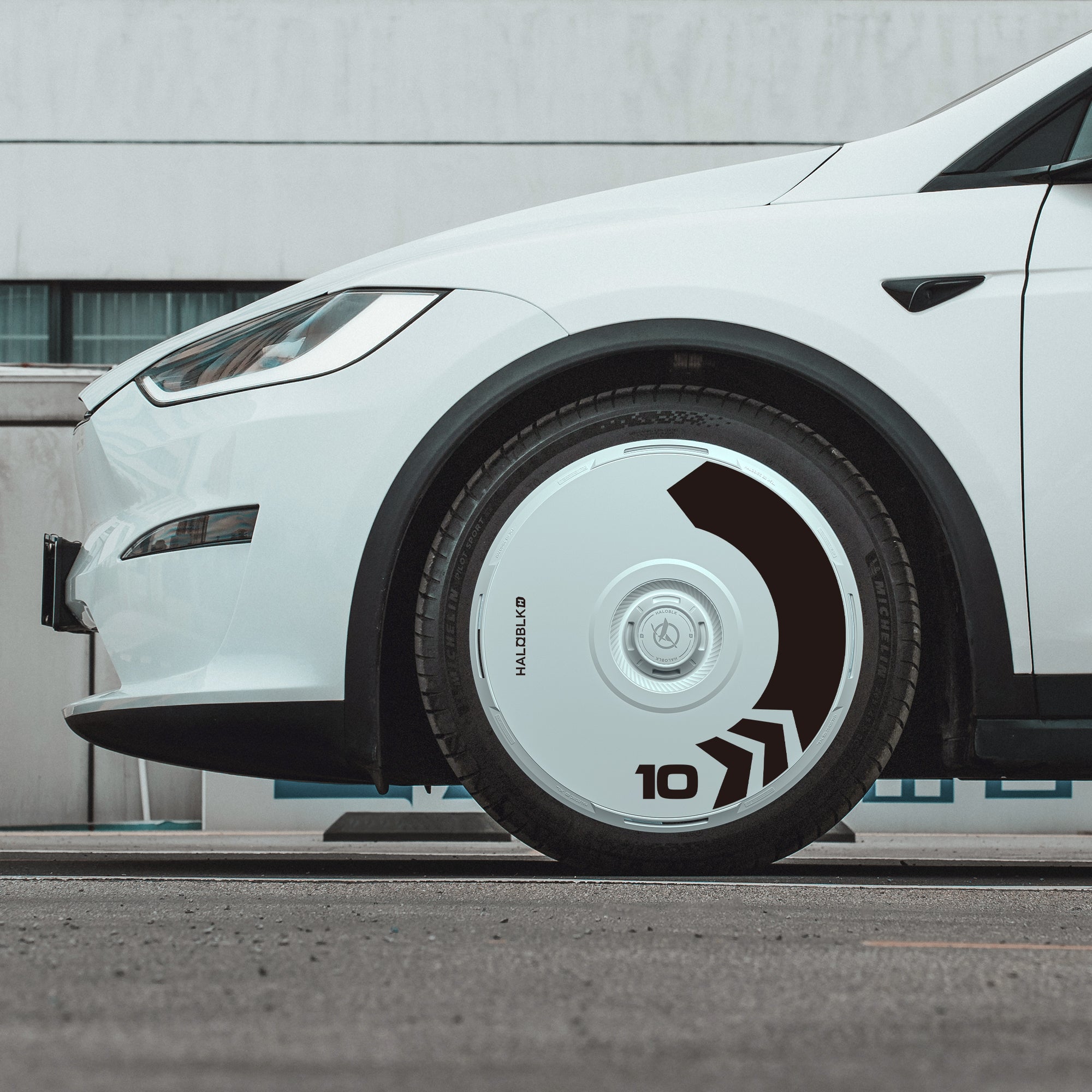 Side view of a white Tesla Model X (2021-2025) parked on a street, equipped with HALODISC 2 Wheel Covers (20" Perihelix Arrow) from HALOBLK Store, featuring bold black graphics, arrows, and the number 10. Gray building with windows in background.