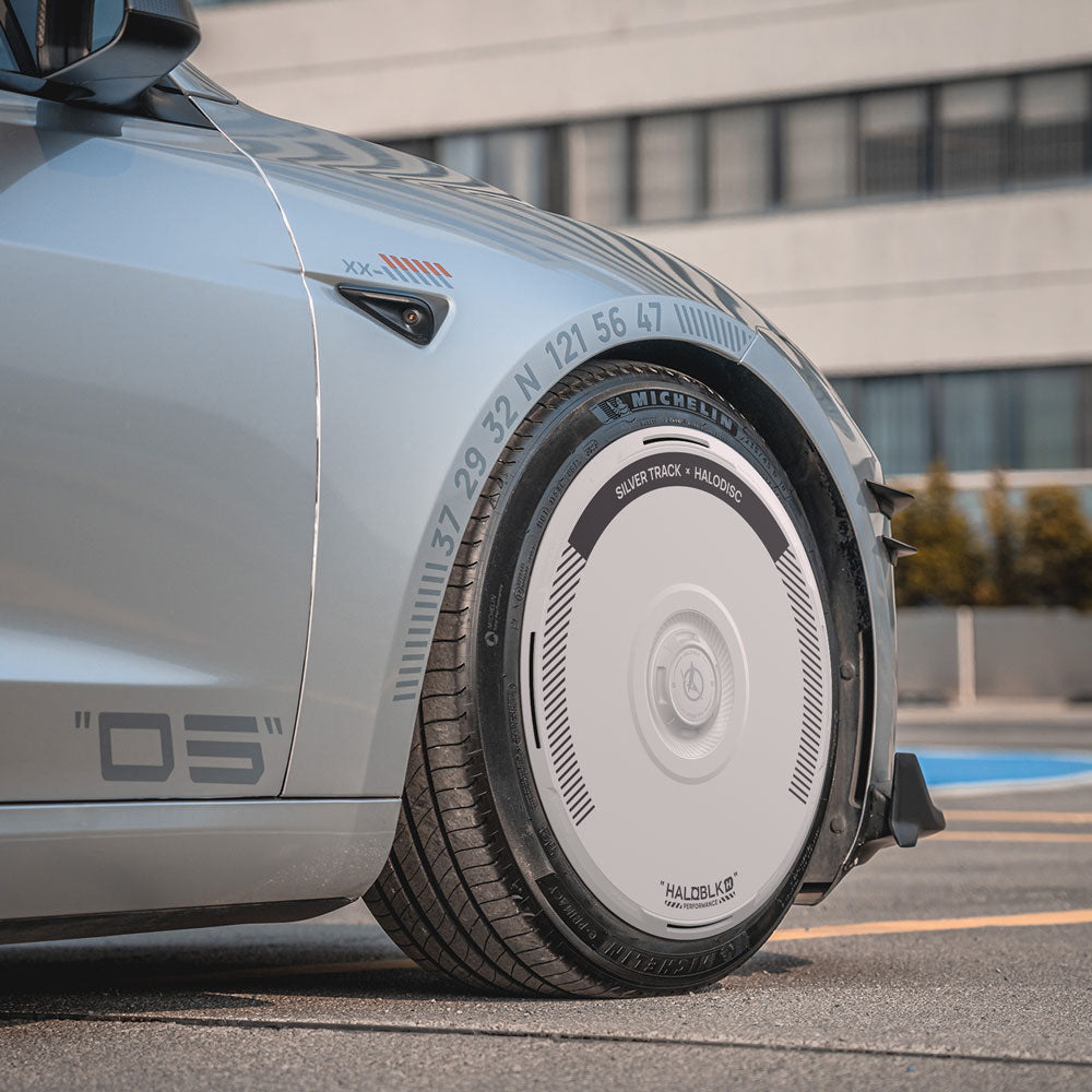 A close-up of a Tesla Model 3 Highland with HALOBLK Store’s HALODISC 2 Silver Track wheel cover (fits 20" Warp Wheels), showing its unique, smooth design with numbers and text; a modern building and greenery appear in the background.