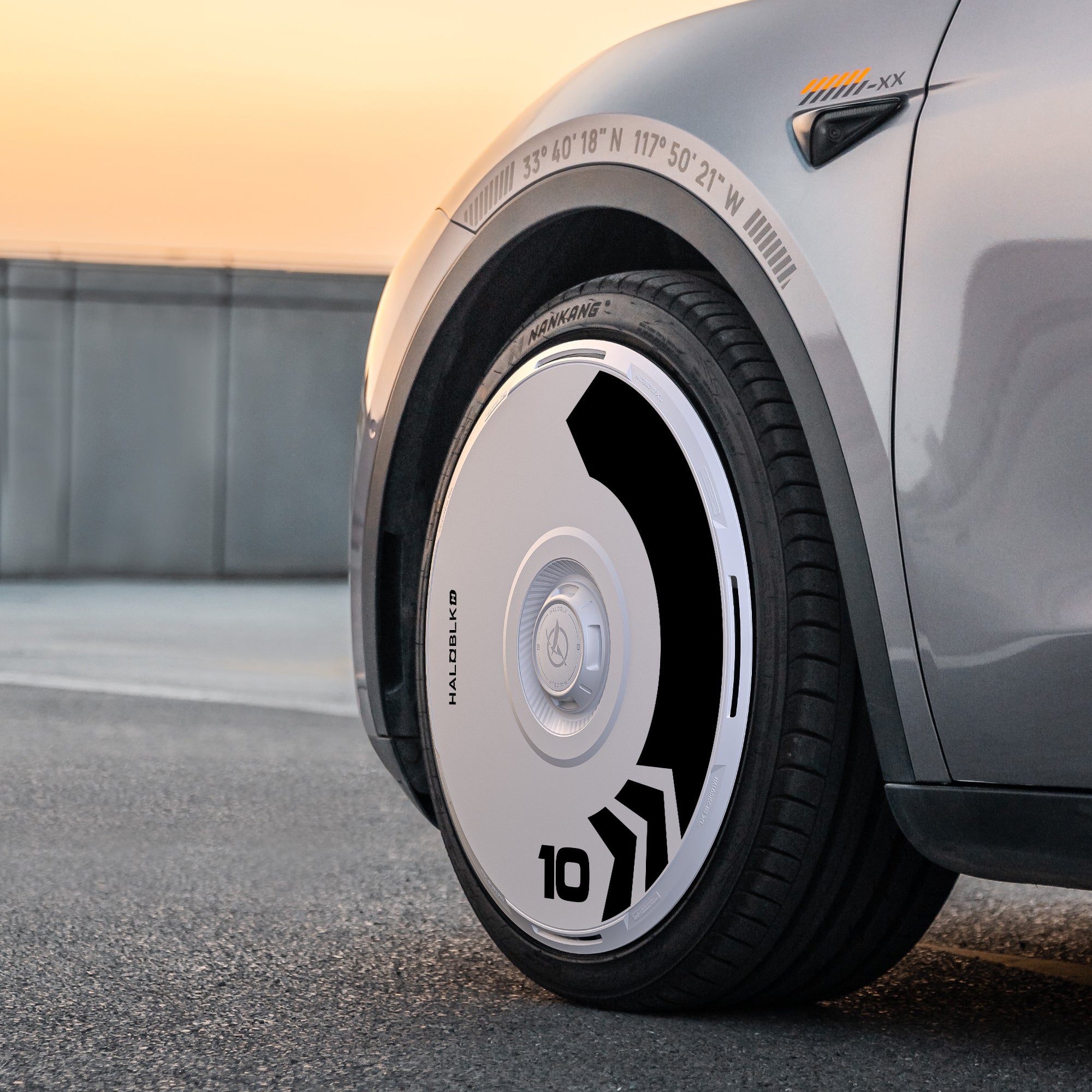 Close-up of a Tesla Model Y’s front wheel with HALODISC 2 Arrow wheel covers by HALOBLK Store, designed in black and white geometric patterns for 20" Induction Wheels. Car is parked on pavement at sunset.