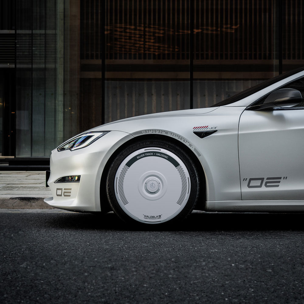 A close-up side view of a white Tesla Model S on a city street, featuring HALODISC 2 Silver Track wheel covers for 21" Arachnid Wheels from HALOBLK Store, with stylized “OE” decals near the front wheel.