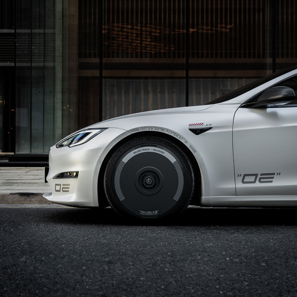 A white Tesla Model S with HALODISC 2 Silver Track wheel covers for 21" Arachnid wheels by HALOBLK Store is parked on a street in front of a modern glass-and-metal building, featuring “OE” decals on its side and front bumper.