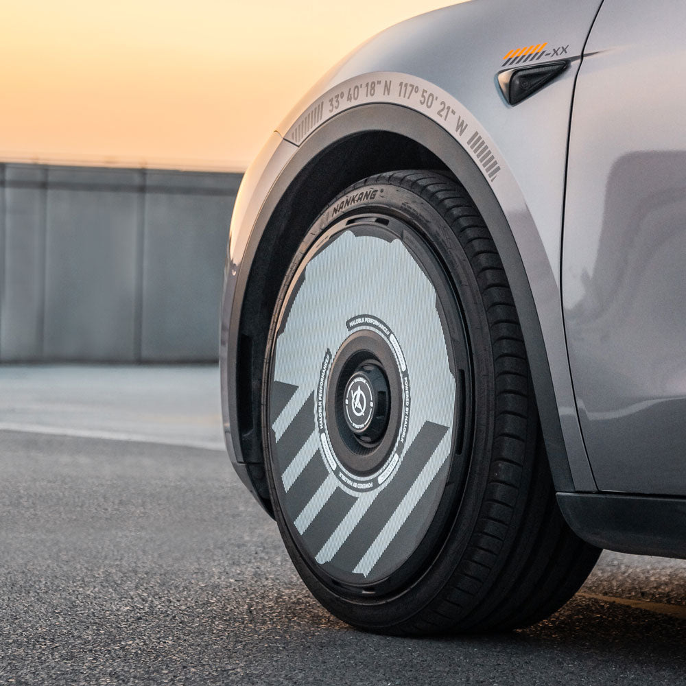 Close-up of HALODISC 2 Wheel Covers for Tesla Model Y 19" Gemini Wheels by HALOBLK Store. The silver car features a modern wheel with a sleek, silver and black aerodynamic cover. Measurements are marked above the wheel arch in a minimal sunset outdoor setting.