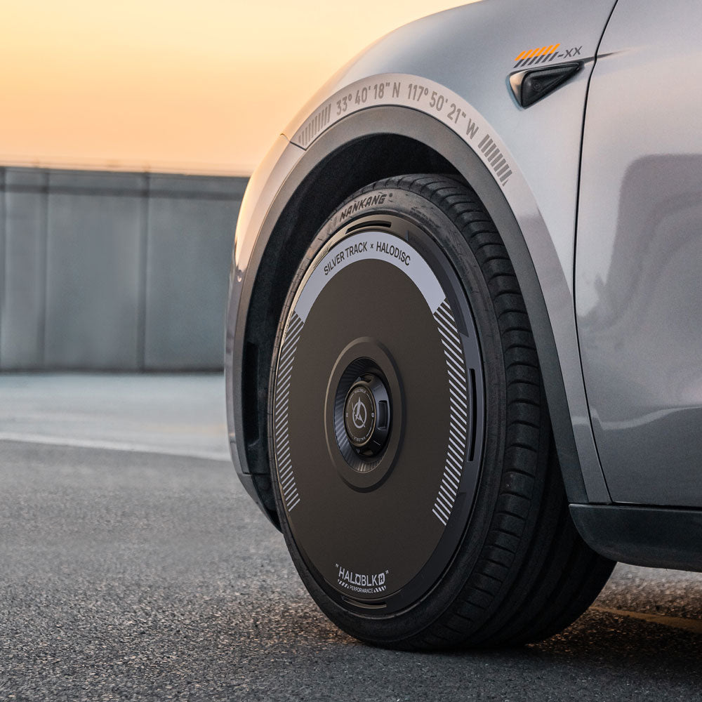 A close-up of a Tesla Model Y’s front wheel with a HALODISC 2 Silver Track cover from HALOBLK Store, designed for 19" Gemini wheels, featuring white markings. The silver car is parked on pavement at sunset, showcasing its modern look.