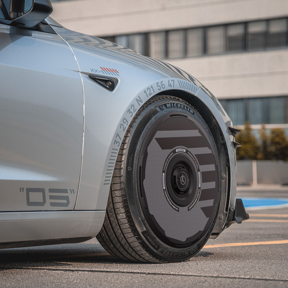 A close-up of HALODISC 2 Wheel Covers for Tesla Model 3 Highland 18" Photon Wheels by HALOBLK Store, showcasing their sleek, futuristic rim design with numeric and line markings, parked near a modern building.