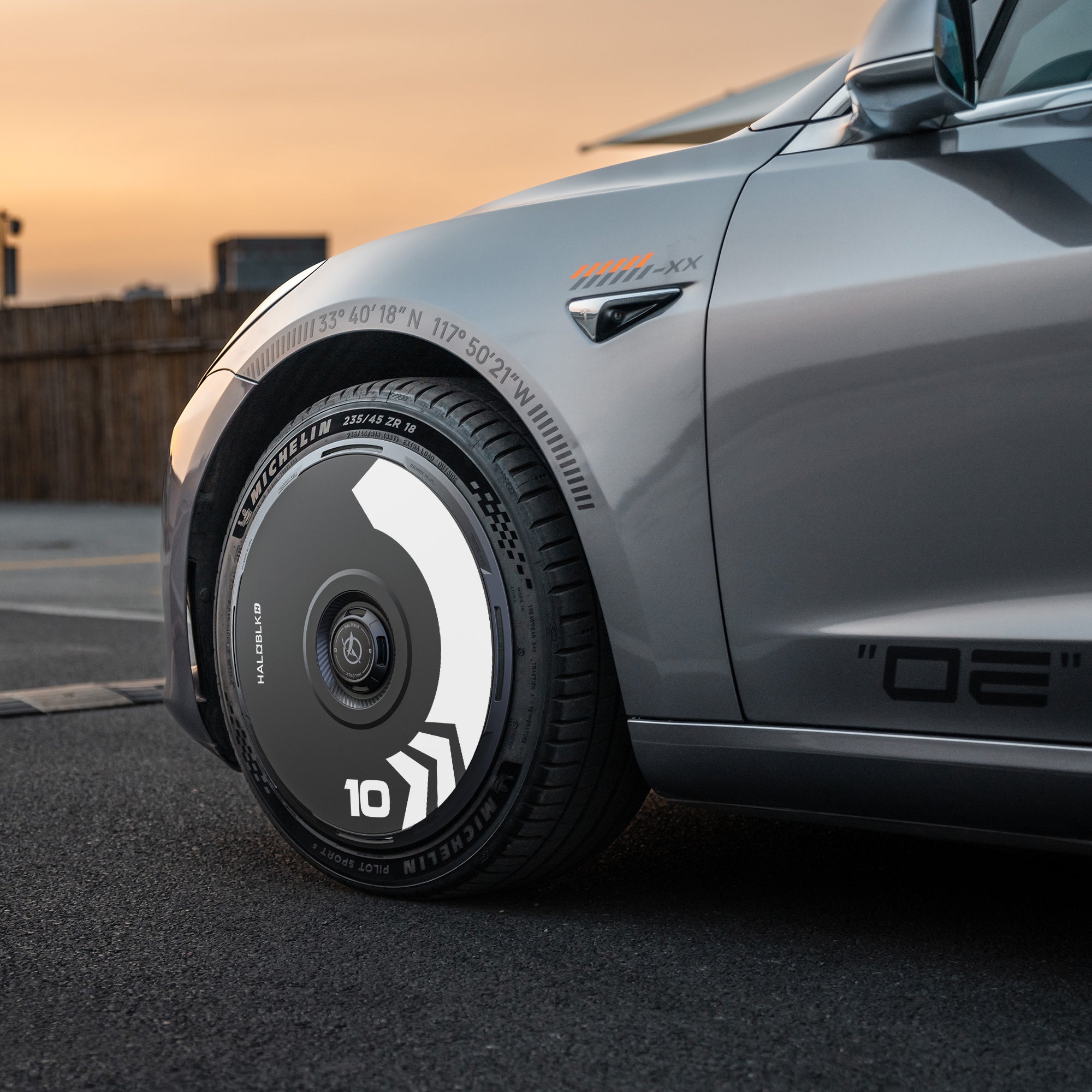 A close-up of a silver Tesla Model 3 (2018-2023) front wheel with HALODISC 2 "Arrow" black and white hubcaps by HALOBLK Store, designed for 19" Stiletto Wheels, featuring chevron stripes, parked on a road at sunset.