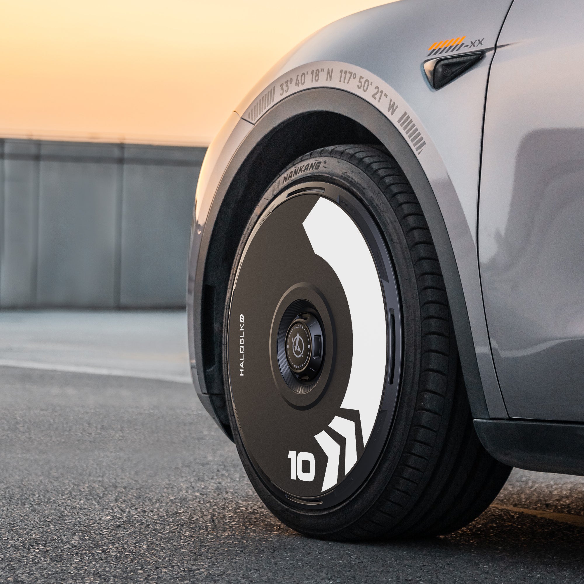 Close-up of HALODISC 2 Wheel Covers for Tesla Model Y 20" Induction Wheels by HALOBLK Store in the Arrow style, showing a black and white futuristic design with bold geometric shapes; part of the car fender and a sunset sky appear in the background.