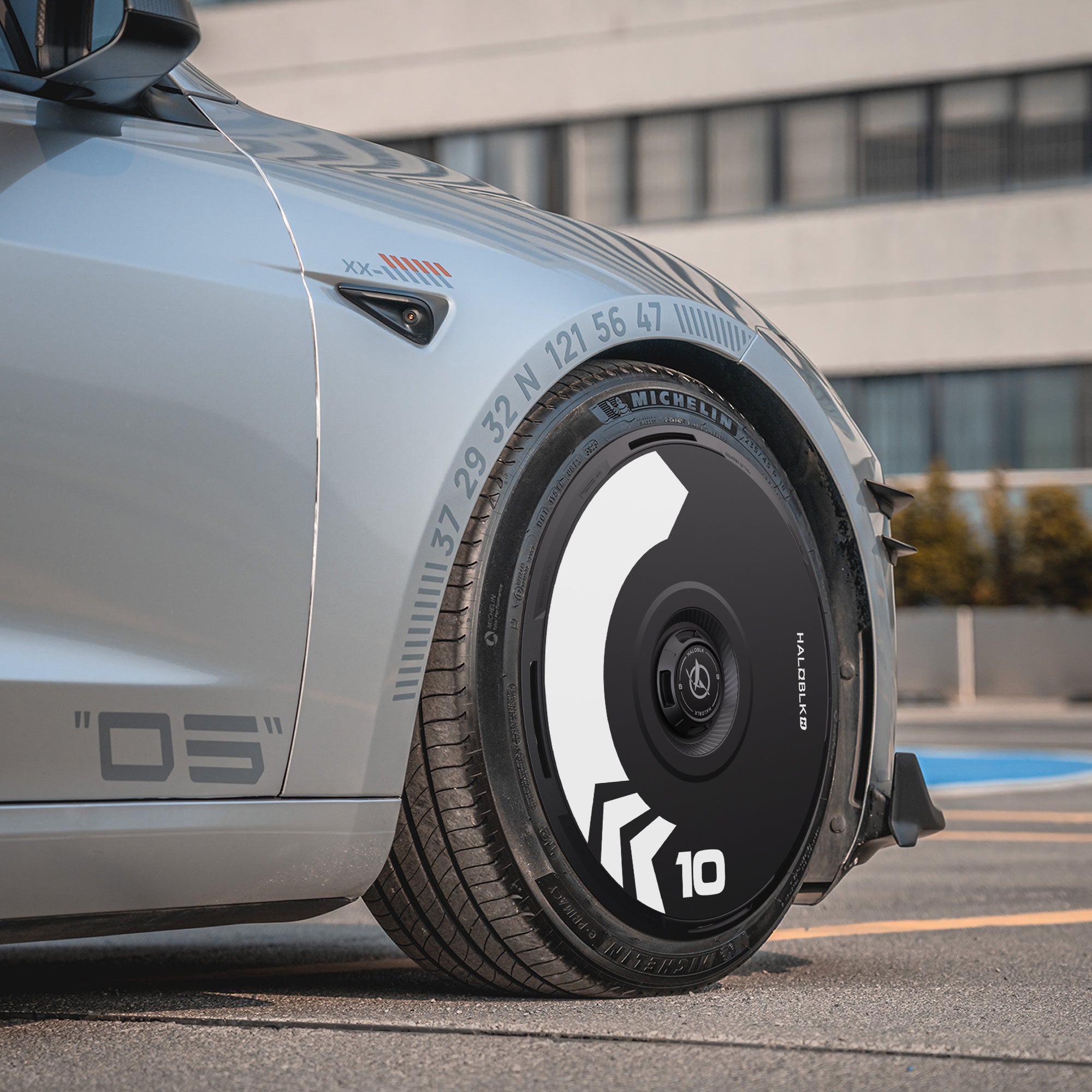 A close-up of a HALODISC 2 Wheel Cover (Arrow) by HALOBLK Store on a silver Tesla Model 3 Highland with 20" Warp Wheels, showcasing its futuristic black and white design and number 10, parked near a modern building.