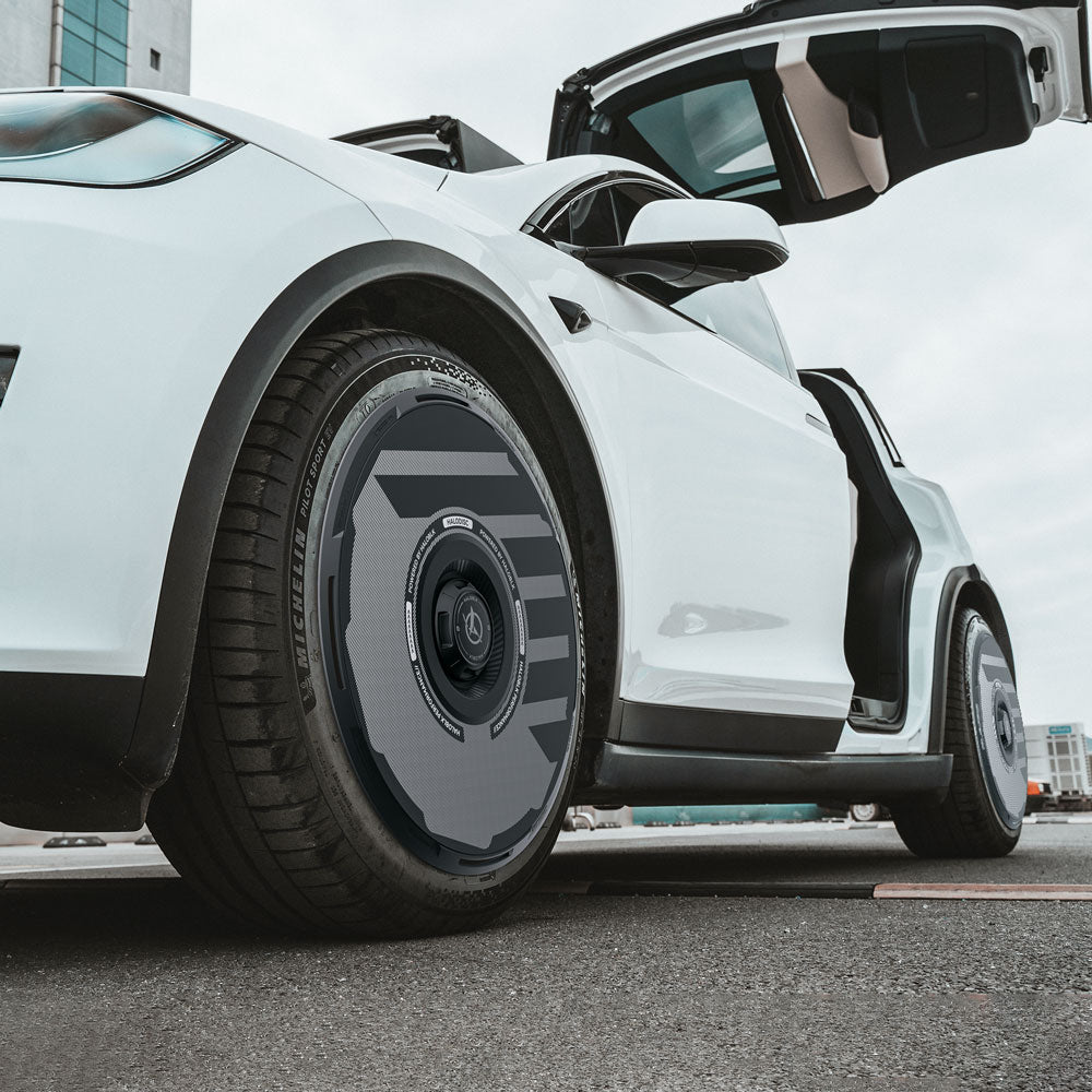 A white Tesla Model X (2021–2025) with gull-wing doors open is parked outdoors, shown from a low angle near the rear wheel, which is fitted with HALODISC 2 black aerodynamic wheel covers by HALOBLK Store for 20" Perihelix Wheels.