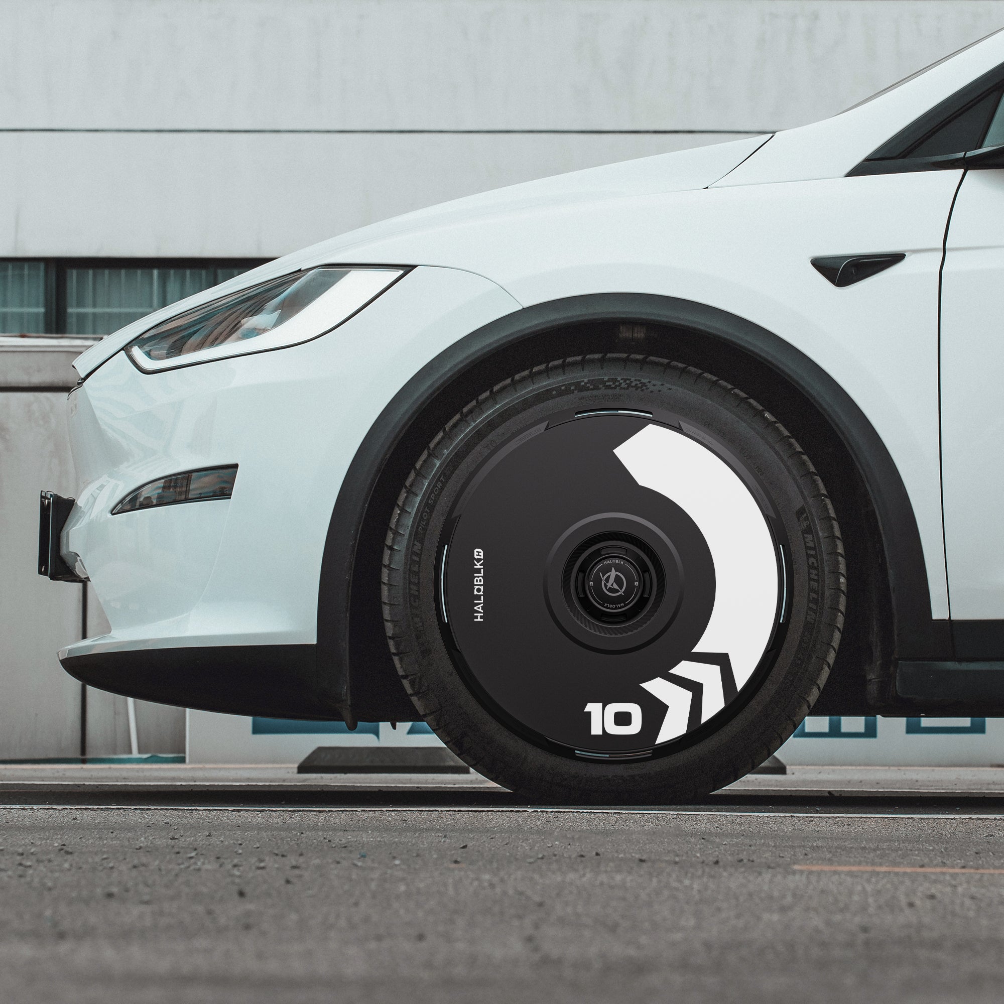 Close-up of a Tesla Model X 2016-2020 front wheel with HALODISC 2 Wheel Covers (Arrow design) by HALOBLK Store—black aerodynamic hubcap with white geometric patterns and the number 10, set against a modern building exterior.