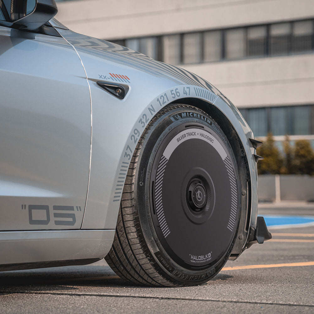 Close-up of a Tesla Model 3 Highland 20" Warp Wheel fitted with HALOBLK Store’s HALODISC 2 Silver Track aerodynamic wheel cover, parked on a paved area near a modern building.