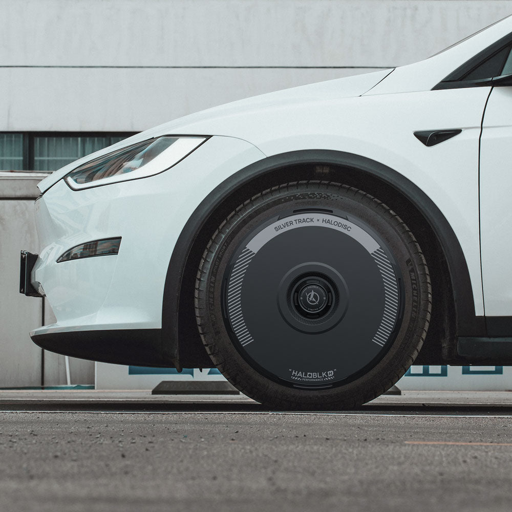 A white Tesla Model X (2021-2025) with 20" Cyberstream wheels is parked outdoors, showcasing a close-up of its HALOBLK Store HALODISC 2 wheel cover in the Silver Track style.