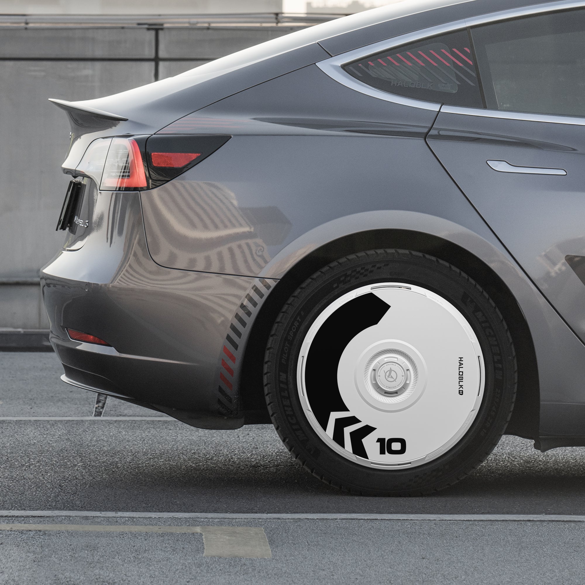 A close-up of the rear of a gray Tesla Model 3 (2019-2023) shows HALODISC 2 "Arrow" 19" wheel covers by HALOBLK Store, featuring futuristic white design with black geometric patterns and the number “10,” parked on a city street.