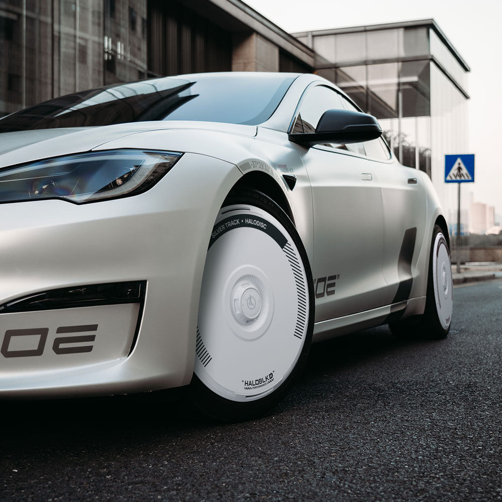 A Tesla Model S with HALODISC 2 Silver Track wheel covers from HALOBLK Store is parked on a city street near a modern glass building and a pedestrian crossing sign.